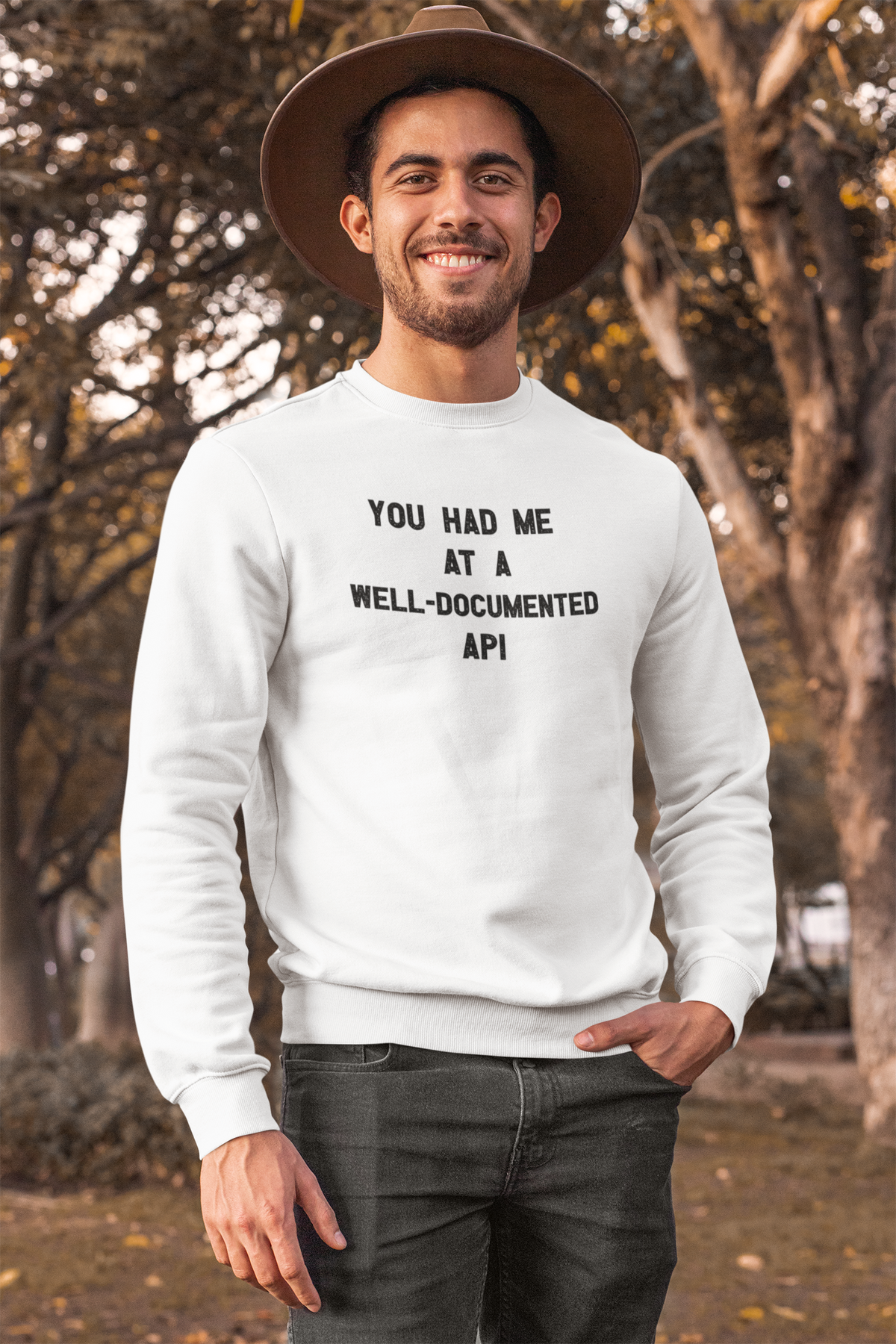 "You Had Me At A Well-Documented API" sweatshirt in white, man in a hat wearing it outdoors, funny developer gift