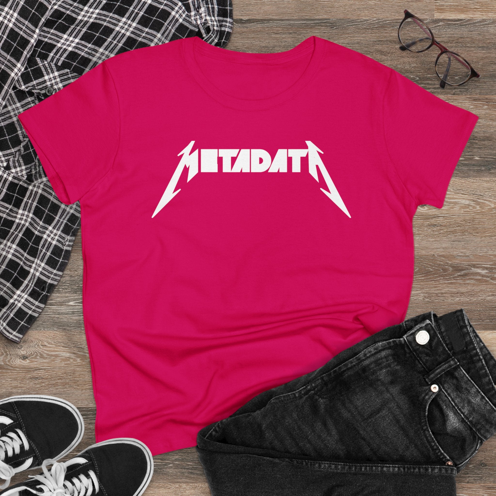The Metadata - Women's Tee is a pink, semi-fitted silhouette made from soft cotton, featuring "METADATA" in a Metallica-inspired font. It adds an edgy touch to your ensemble and is creatively displayed on a wooden floor next to a plaid shirt, black jeans, glasses, and sneakers.