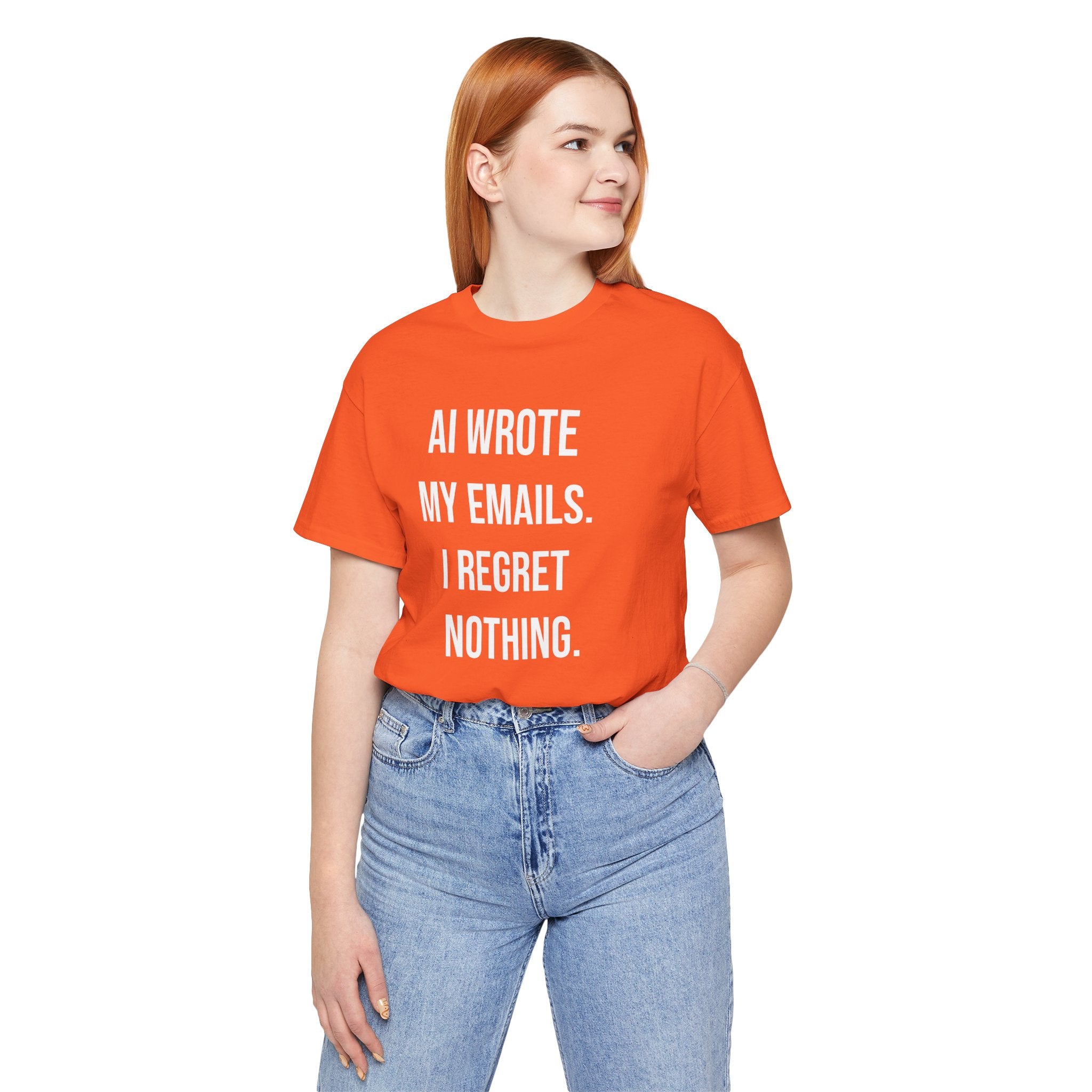 A red-haired woman models the "AI WROTE MY EMAILS I REGRET NOTHING - T Shirt" in bright orange, her hand in her jeans pocket as she glances aside, showcasing this fun programmer humor tee.