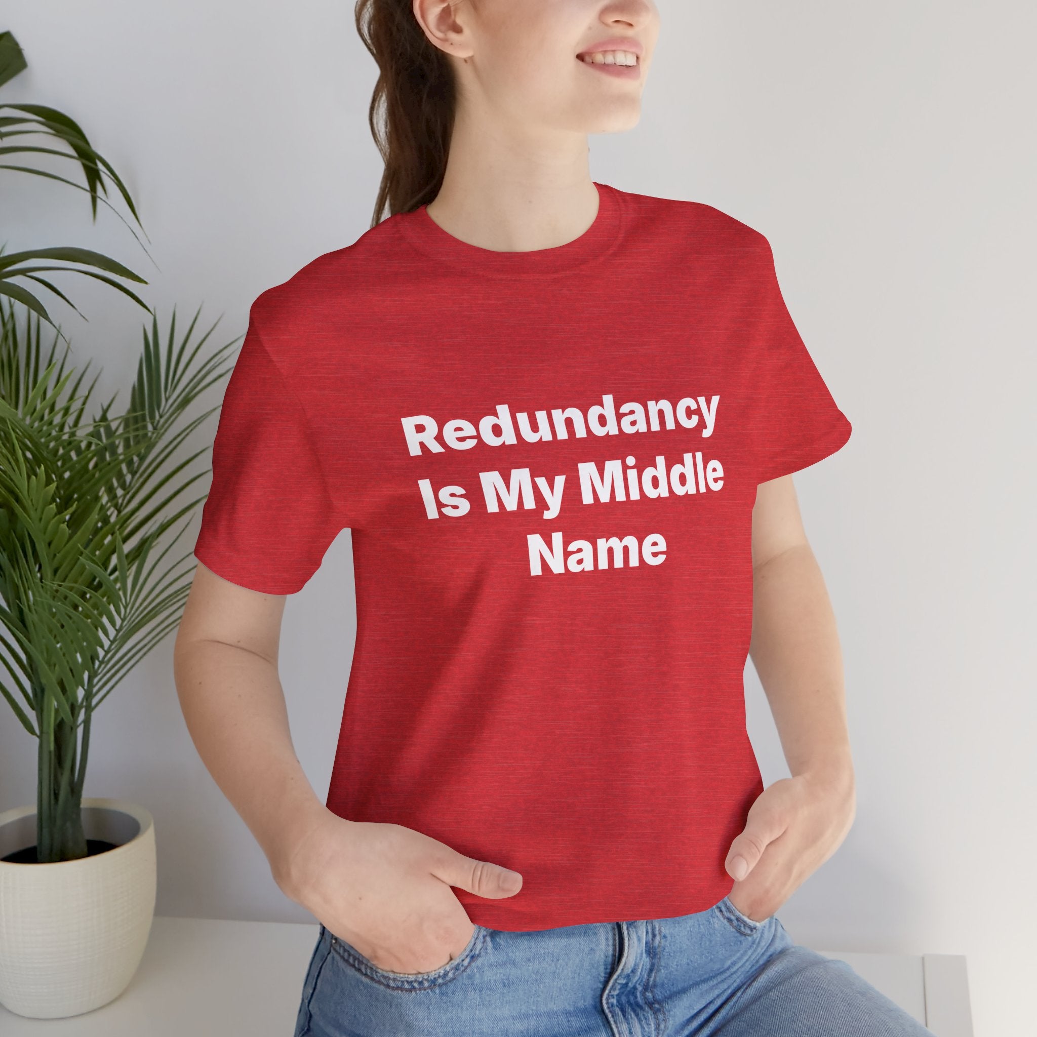 A person wearing a vibrant "Redundancy Is My Middle Name" T-shirt, made from soft ring-spun cotton, is standing next to a plant.