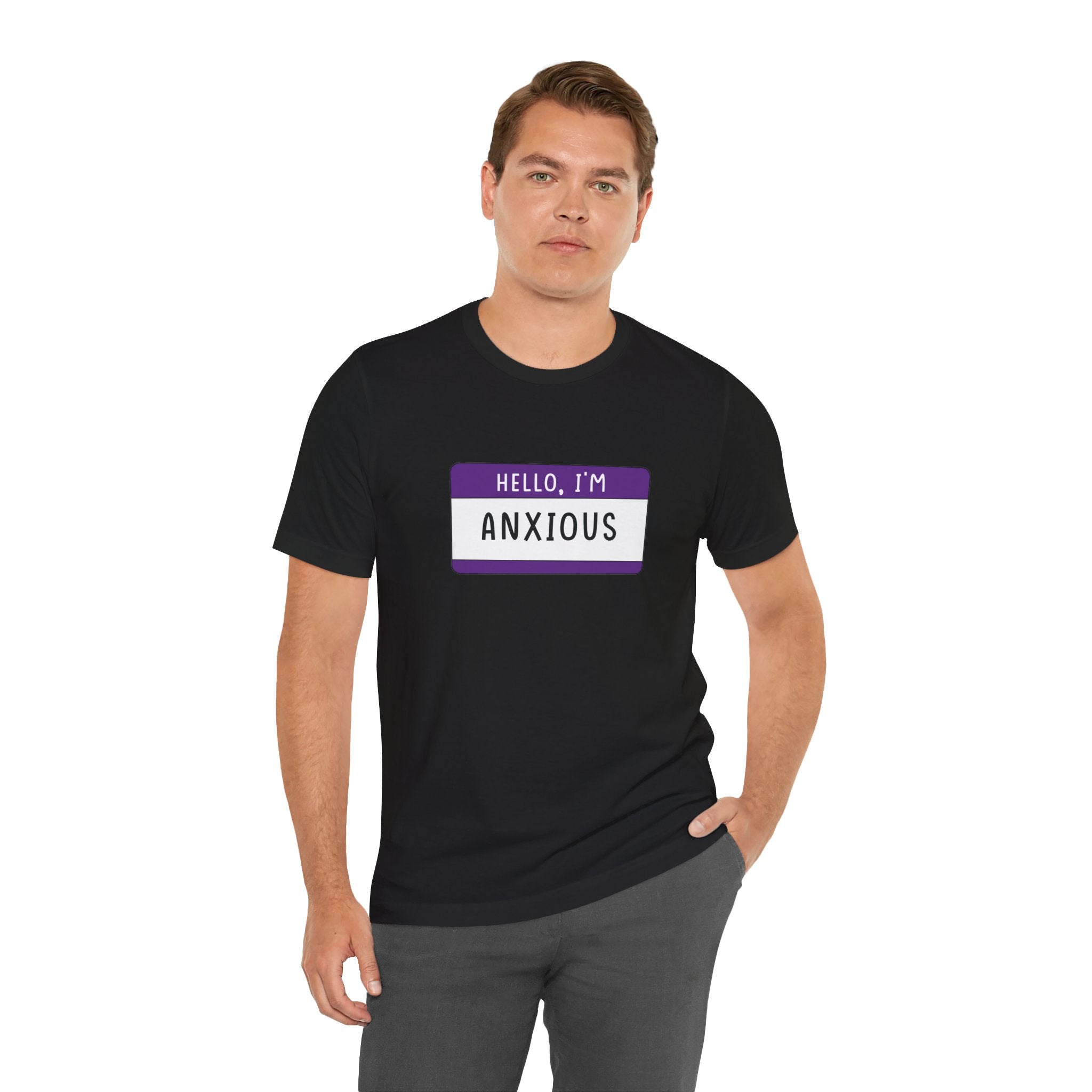Wearing a "Hello, I'm Anxious" T-shirt in black featuring a playful name tag design, an individual stands against a white backdrop. This shirt is ideal for those who enjoy whimsical designs or are geeks and gamers at heart.