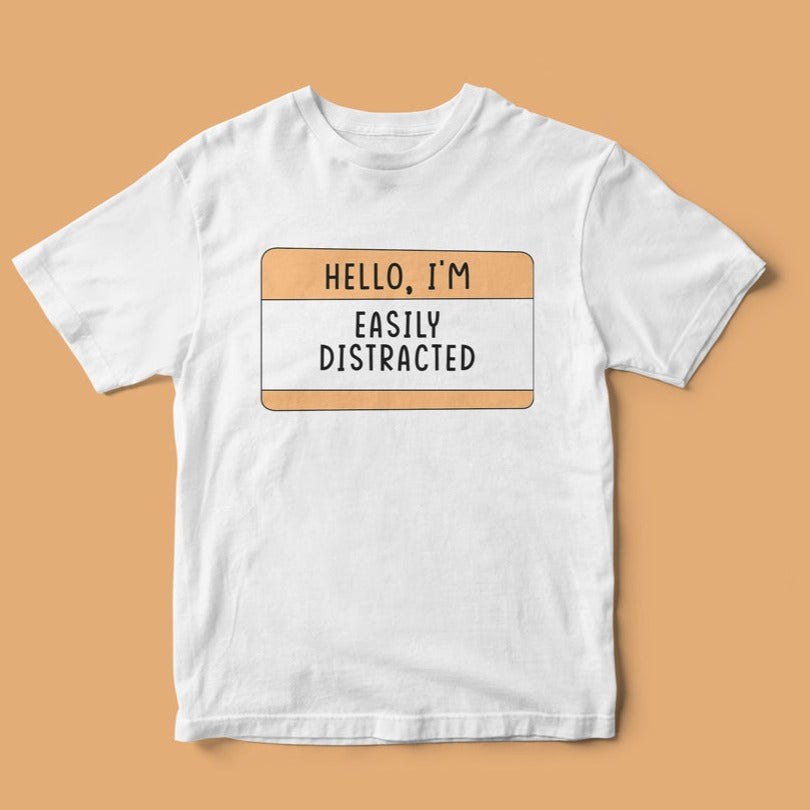 The "Hello, I'm Easily Distracted" T-shirt features a geeky humor design with a name tag that states "HELLO, I'M EASILY DISTRACTED" in bold black letters on a solid beige background.