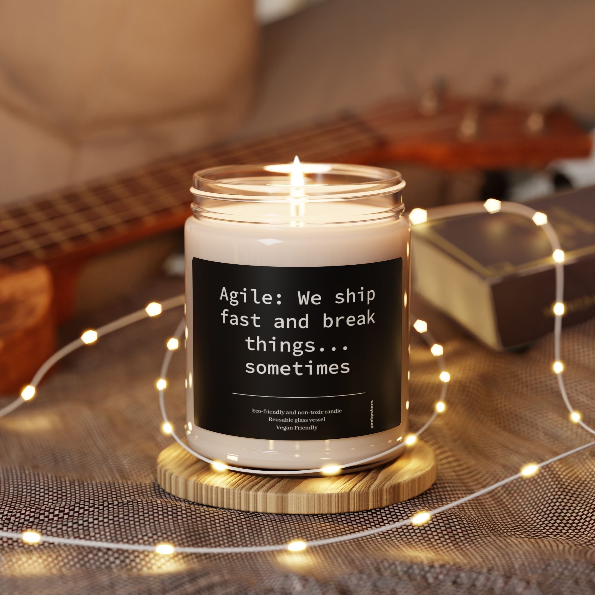 The "Agile: We Ship Fast and Brake Things" candle, an eco-friendly soy blend, emits a warm glow. It's surrounded by cozy string lights with a blurred background featuring a guitar and a book.