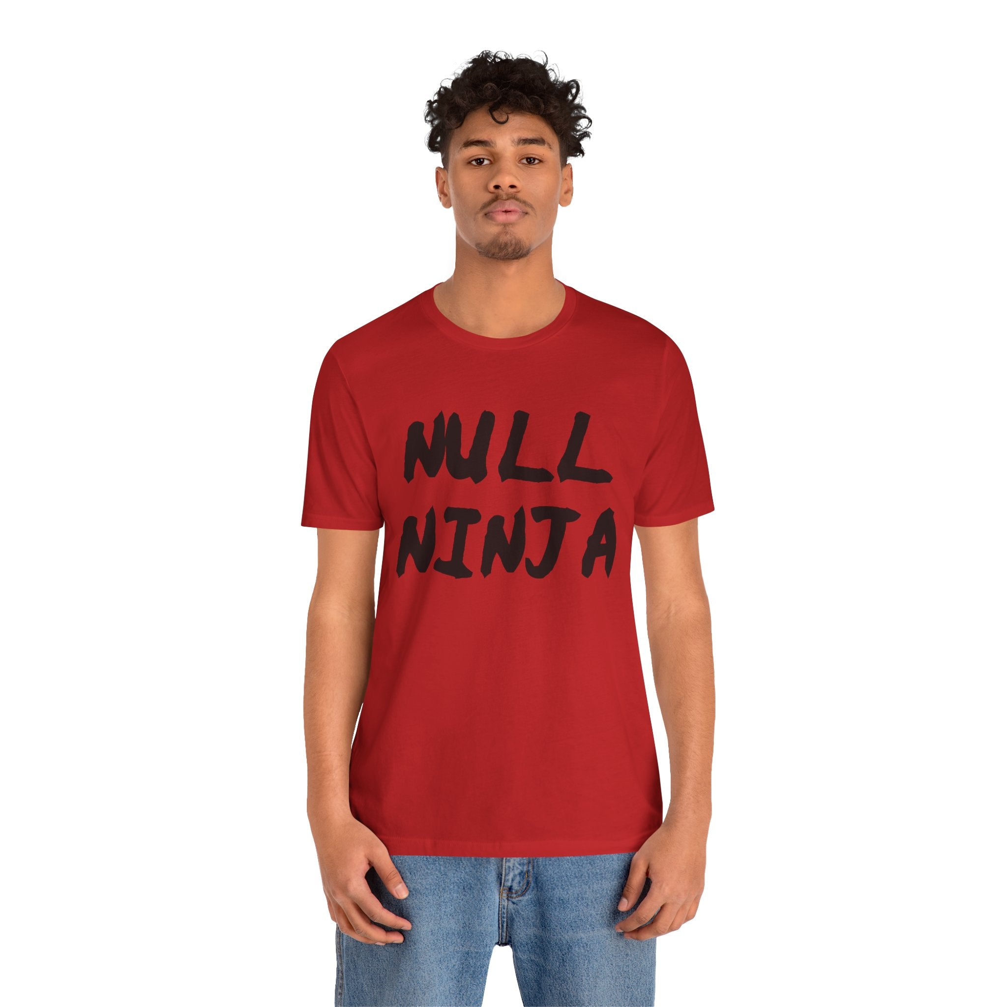 Someone confidently stands against a plain white background wearing the red Null Ninja Tee Shirt, featuring "NULL NINJA" in bold black print, along with blue jeans.