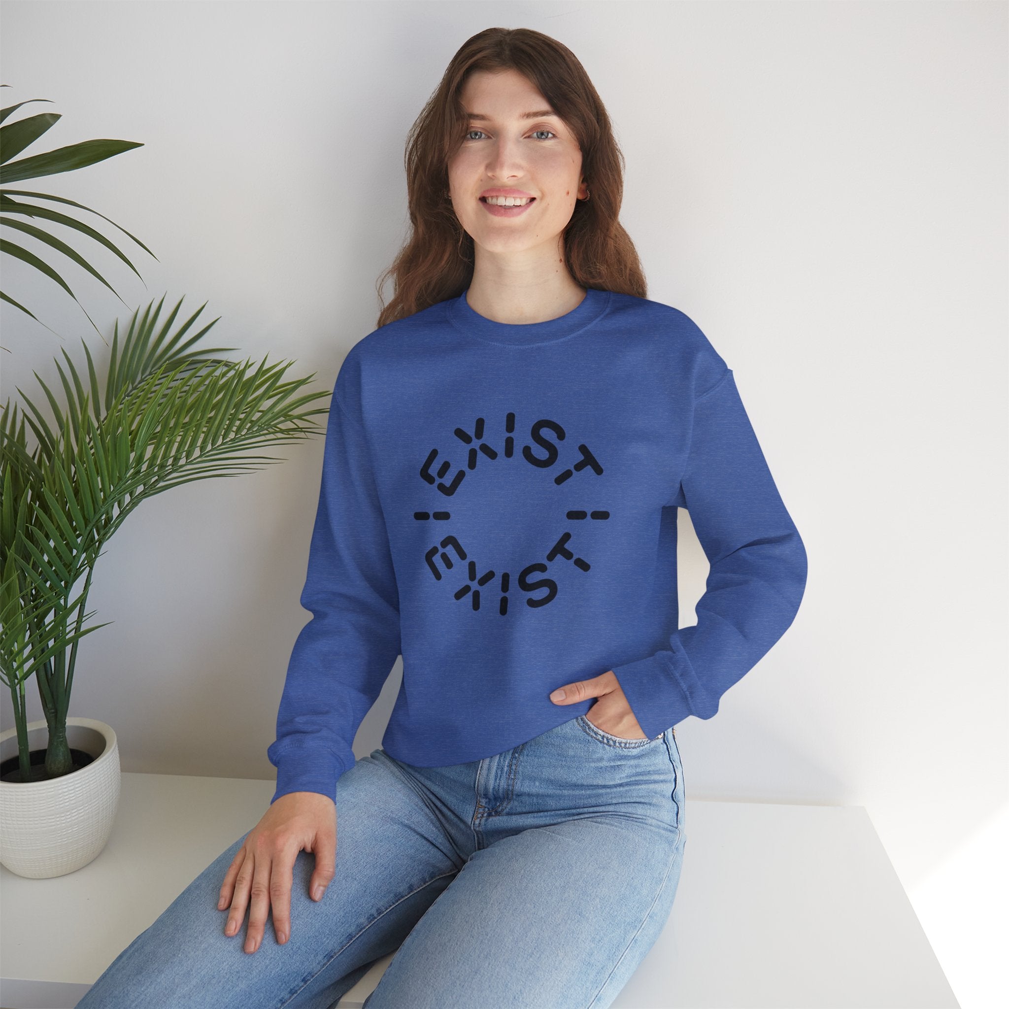 Wearing the eco-friendly Exist Exist - Sweatshirt in blue, featuring the word "EXIST" in a circular pattern, a person sits on a white surface beside a potted plant.