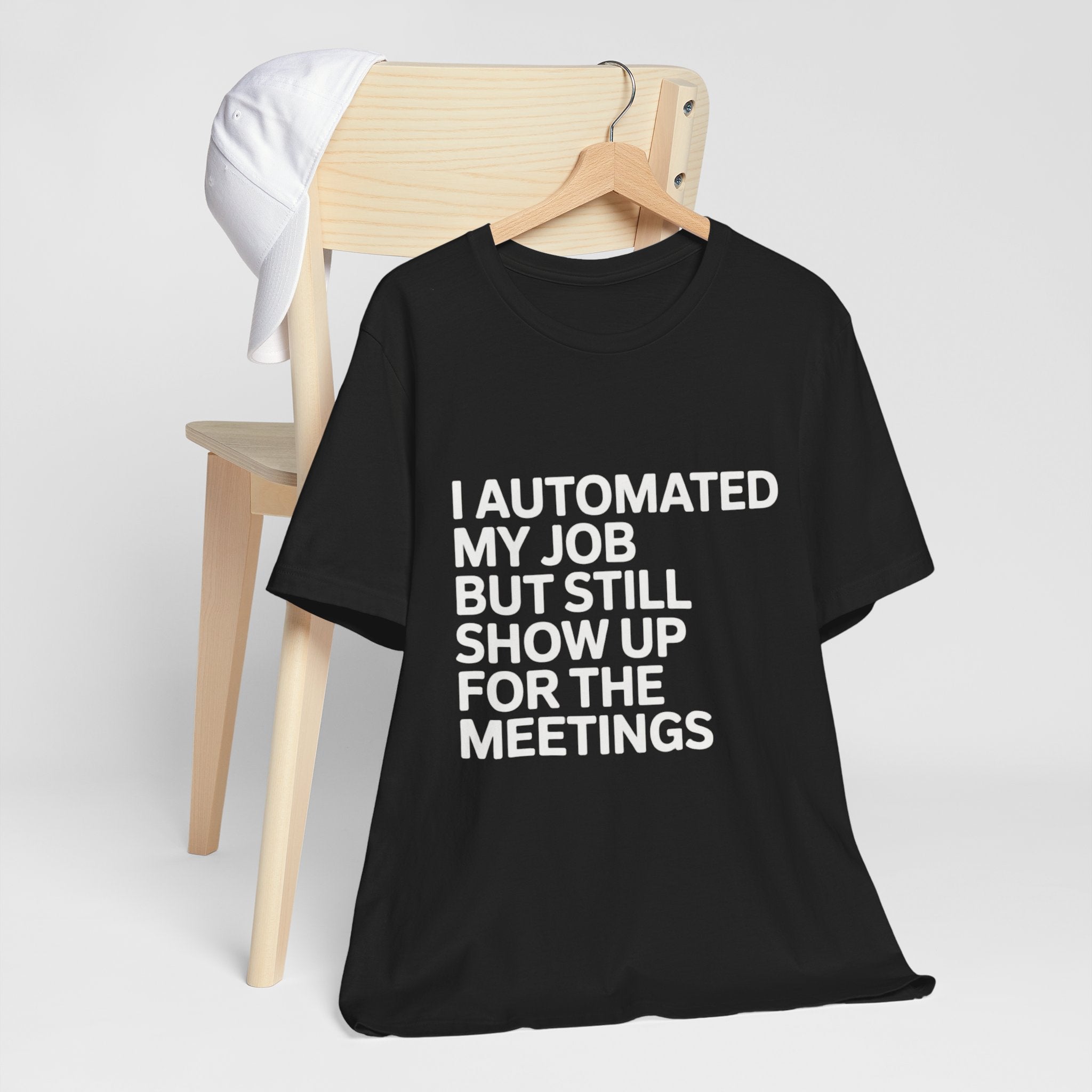 The black "I Automated My Job But Still Show Up for the Meetings" t-shirt hangs on a hanger over a wooden chair, paired with a white cap.