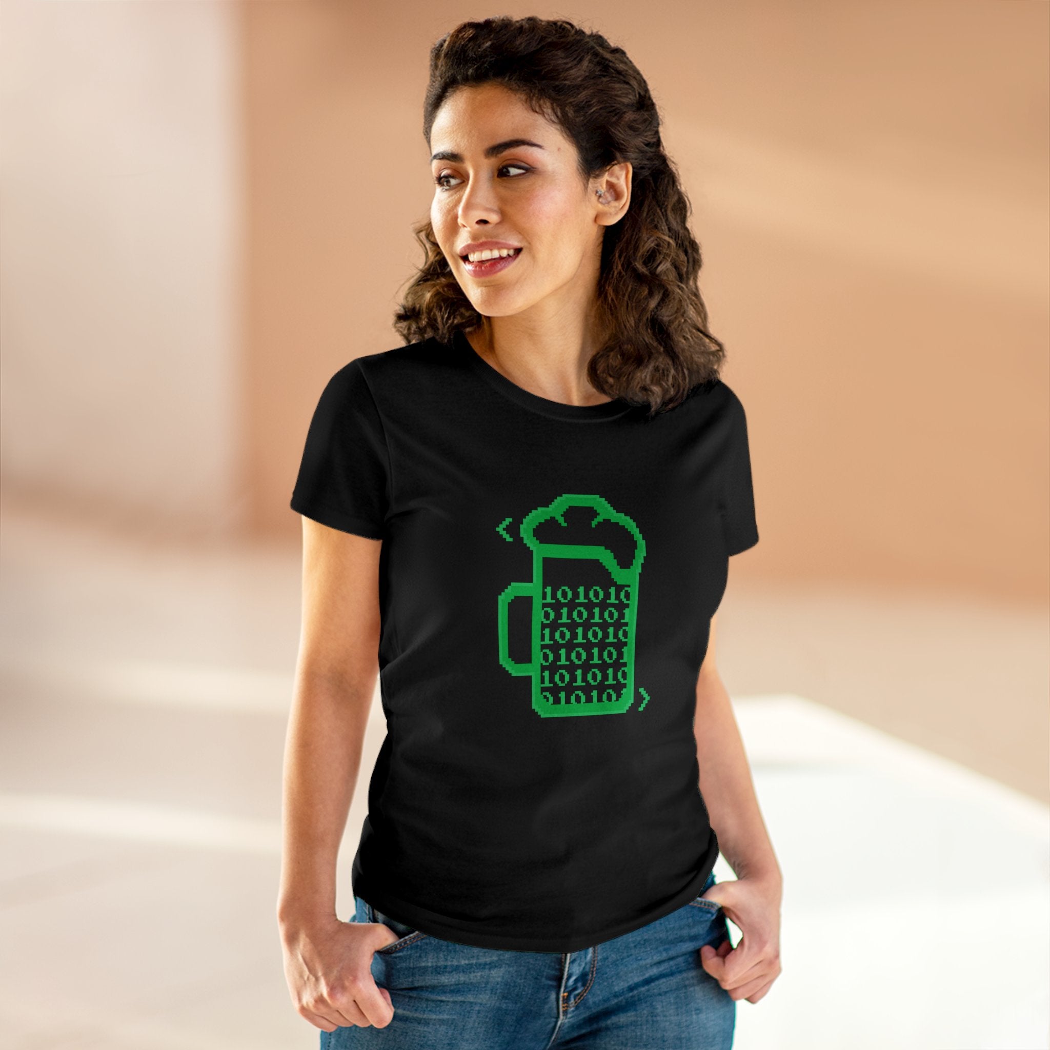 A woman stands indoors, wearing the Beer Code - Women's Tee— a black shirt with a coffee cup and binary code design—looking to the side and proudly embracing geek culture.