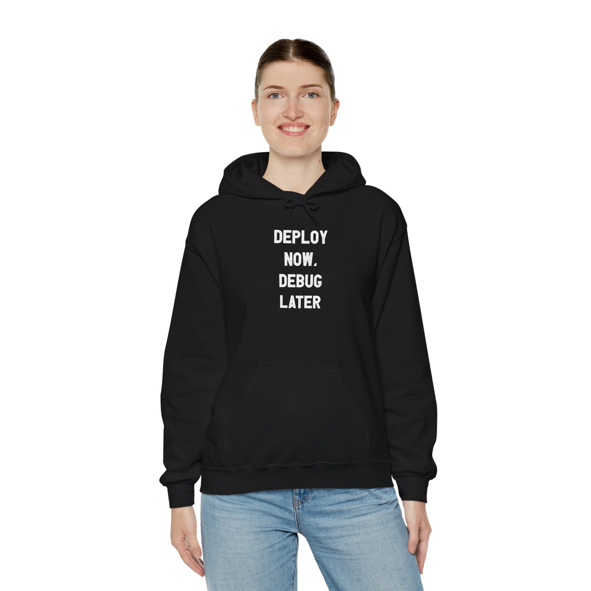 A person wearing a "Deploy Now, Debug Later" Hooded Sweatshirt paired with jeans exudes comfort and style while standing against a white background.