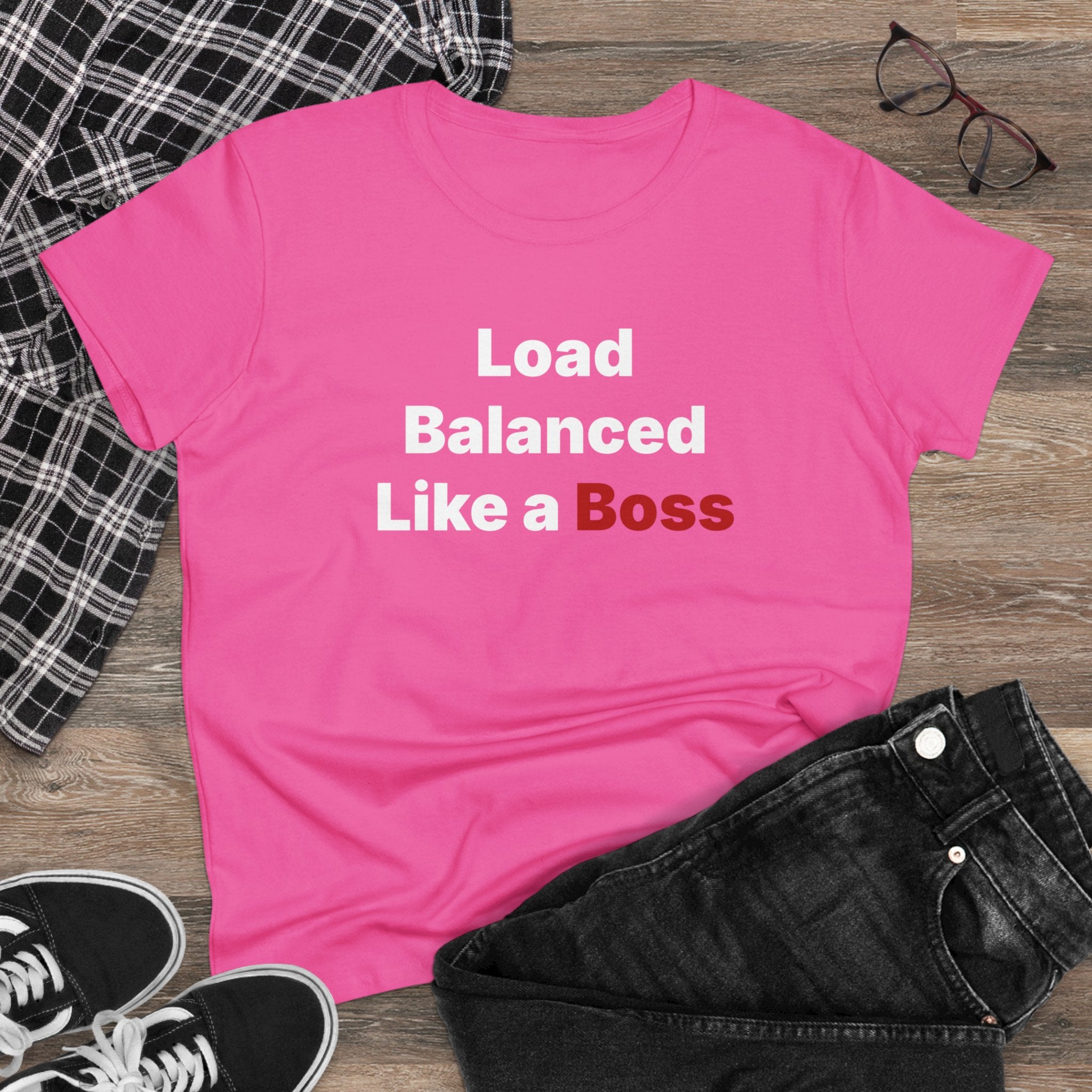 The "Load Balanced Like a Boss" women's tee in pink features cap sleeves and displays text in white and red. It's styled on a wooden surface with a black plaid shirt, black jeans, black sneakers, and glasses.