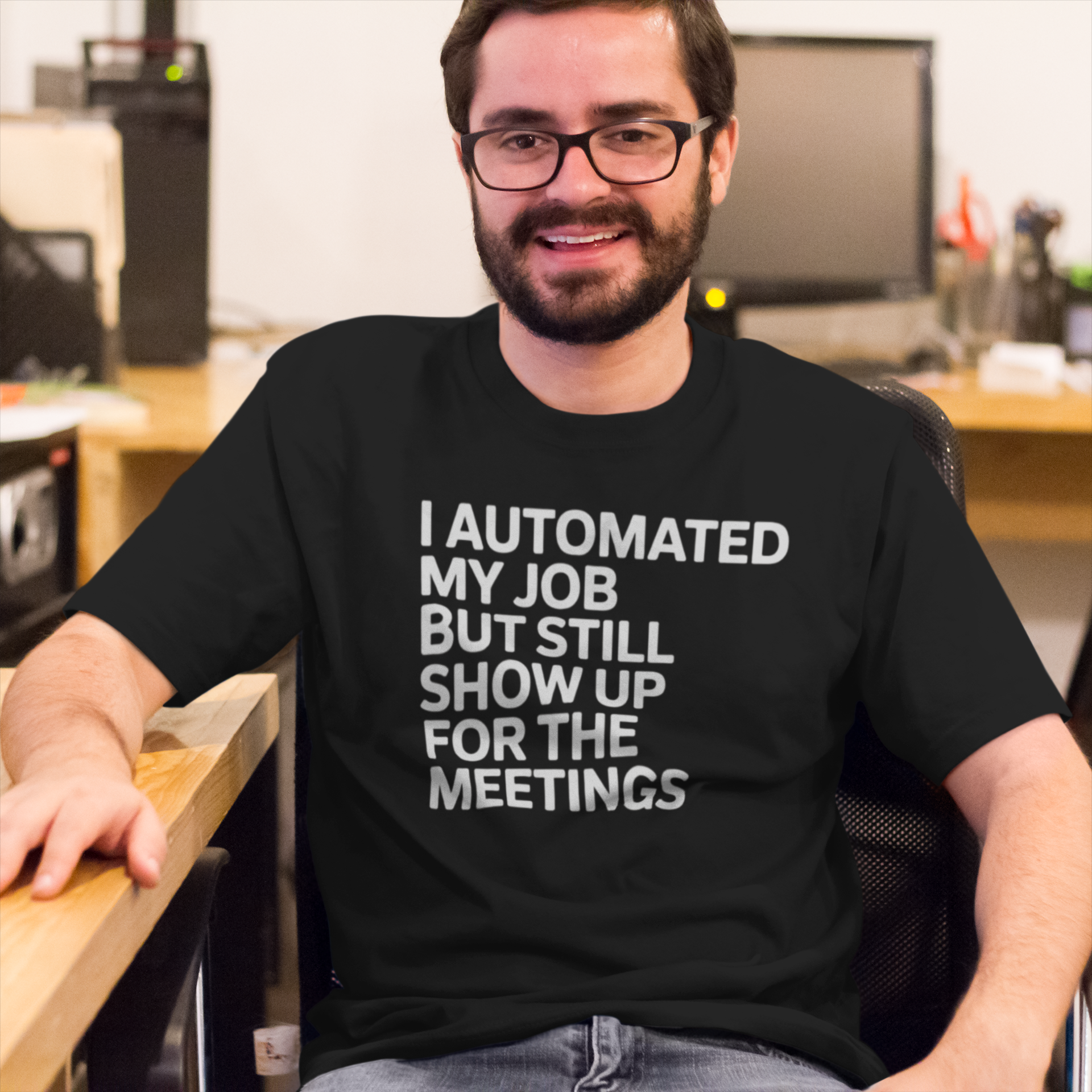 A man sits at his office desk wearing the "I Automated My Job But Still Show Up for the Meetings" tech humor shirt.