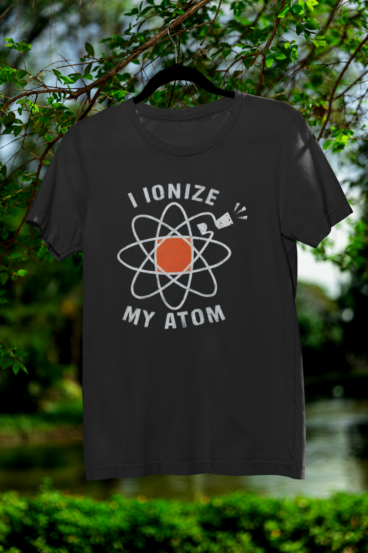 'I Ionize My Atom' t-shirt in black with atom icon print, hanging outdoors on tree, funny science gift