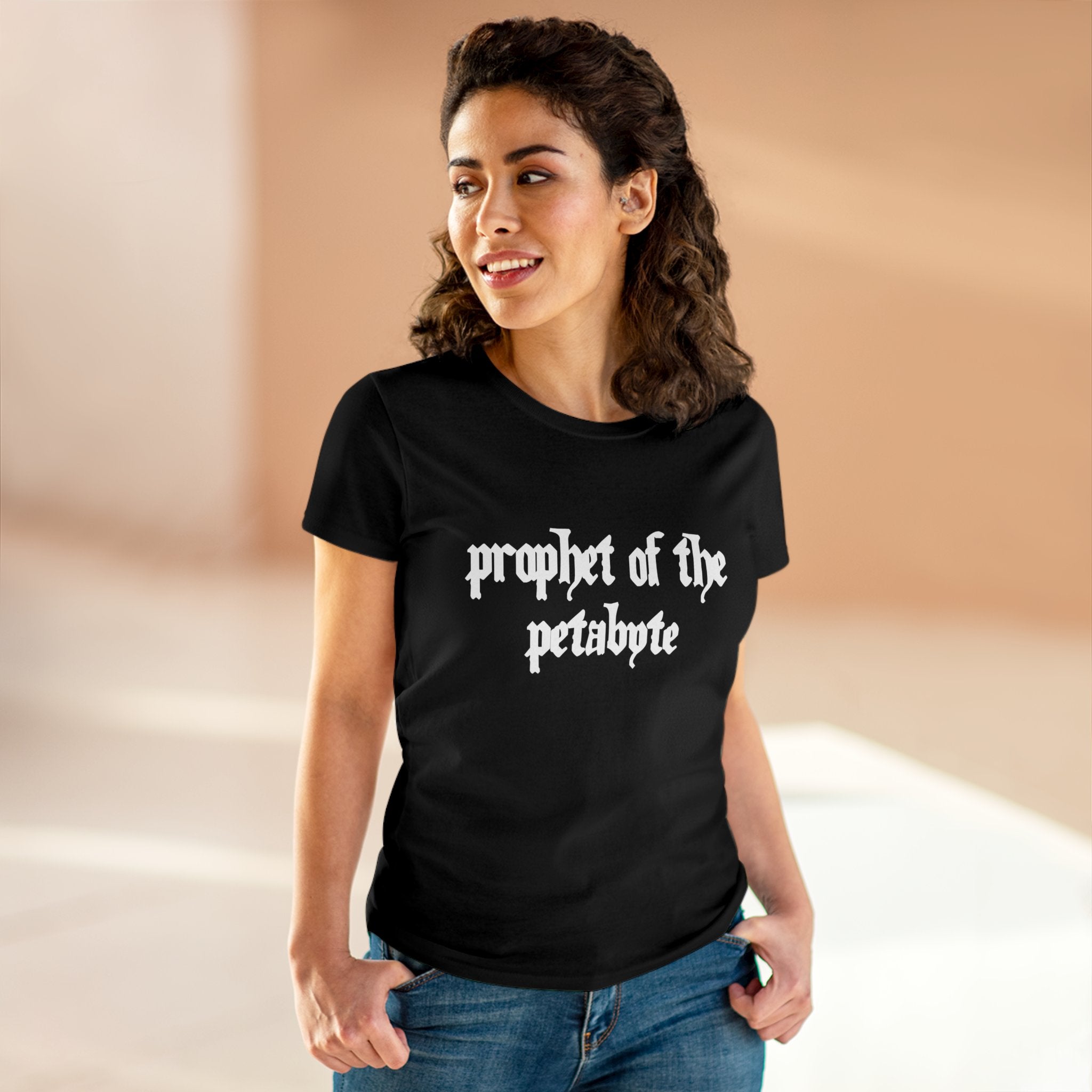 A person with curly hair is wearing the "Prophet of the Petabyte - Women's Tee," a black, soft cotton shirt, while standing indoors.