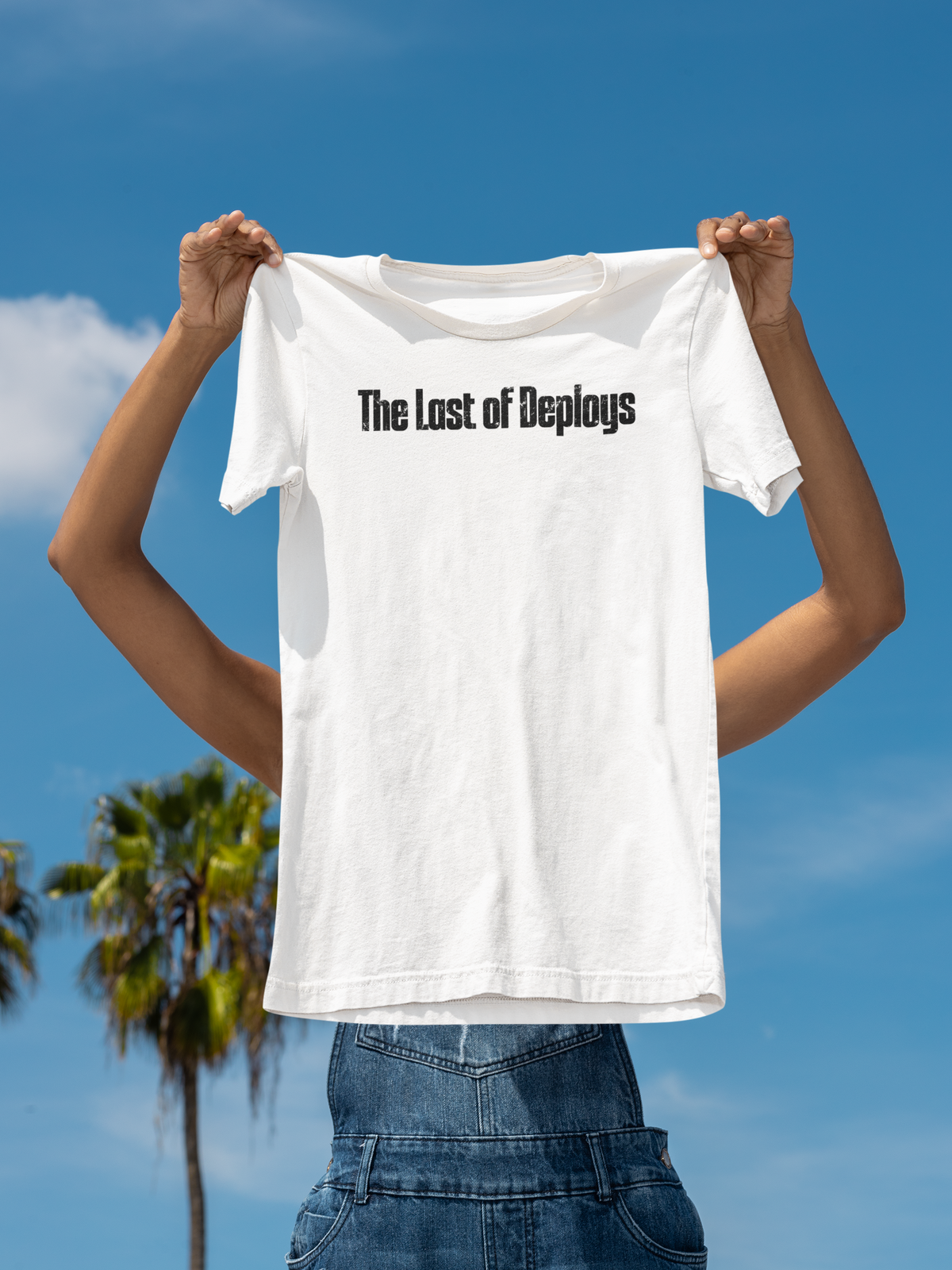 'The Last of Deploys' T-shirt in white, woman holding it up against a blue sky backdrop, gamer gift tee