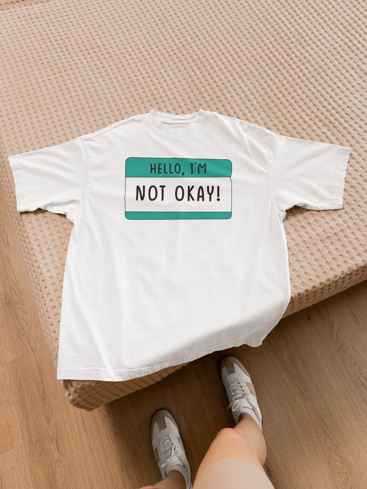 Hello I'm Not Okay name tag T-shirt in white, flat lay on wood floor with shoes, sarcastic gift tee