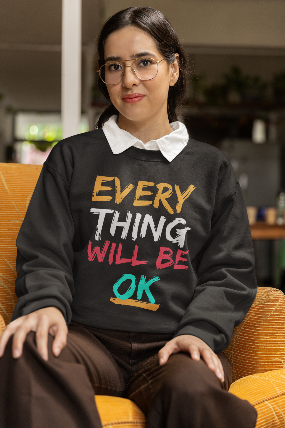 'Everything Will Be OK' sweatshirt in black, woman sitting indoors wearing it, uplifting gift