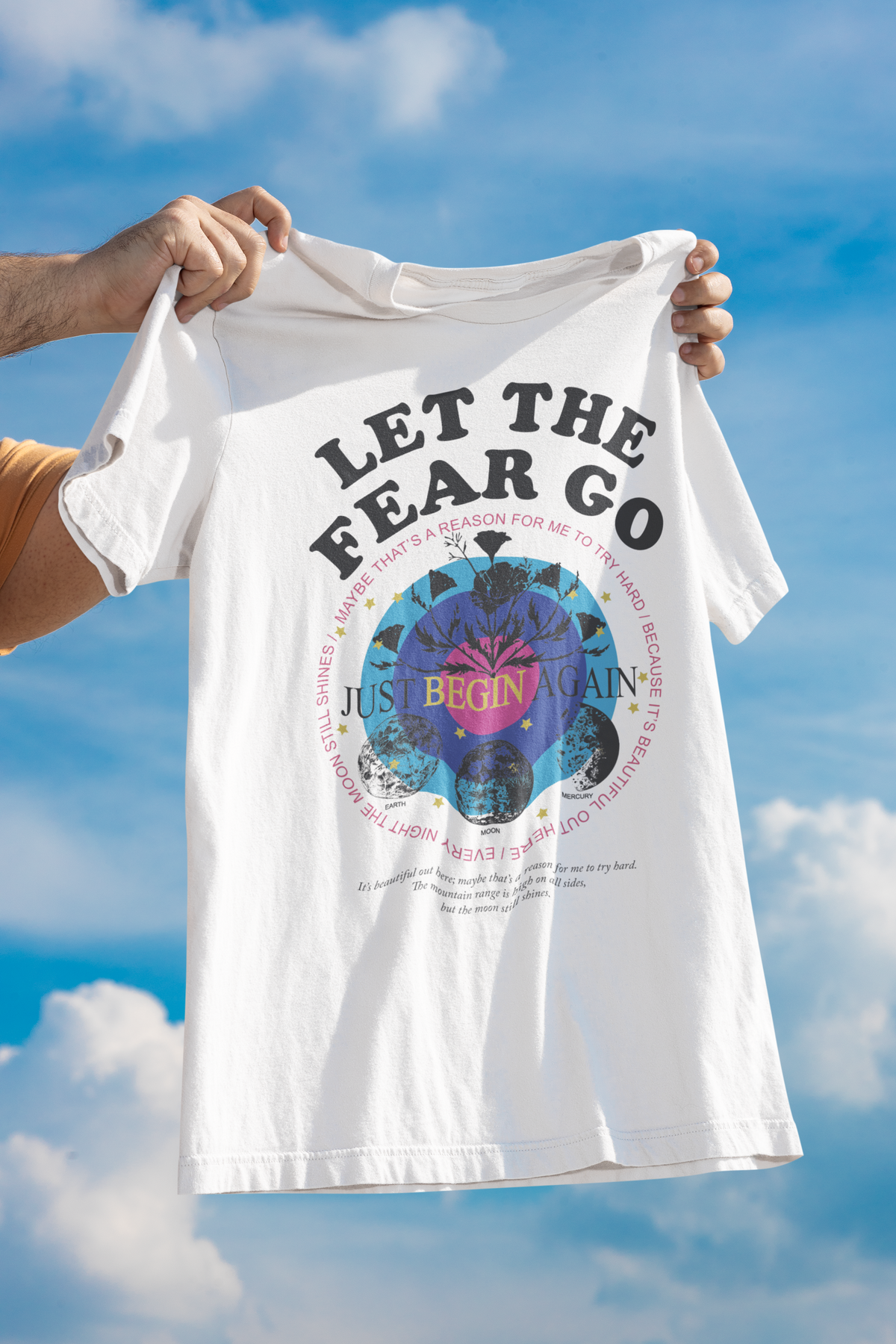 "Let the Fear Go" cosmic graphic t-shirt in white, held up outdoors against sky, inspirational gift
