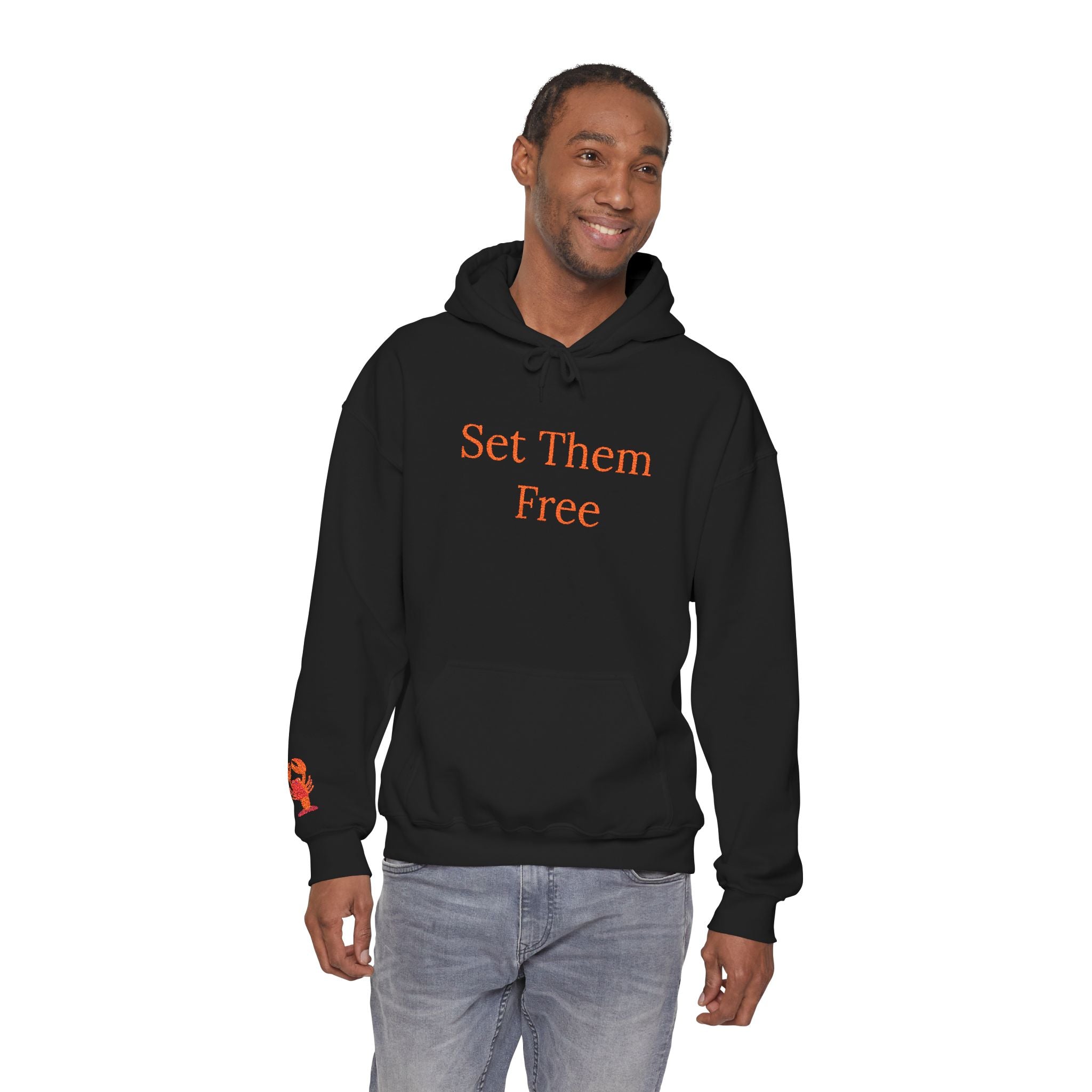 A man wearing the Set Them Free Hoodie — Claude / OpenClaw Moltbot AI Graphic Hoodie in black, featuring "Set Them Free" in orange, paired with light blue jeans, stands and smiles against a white background.