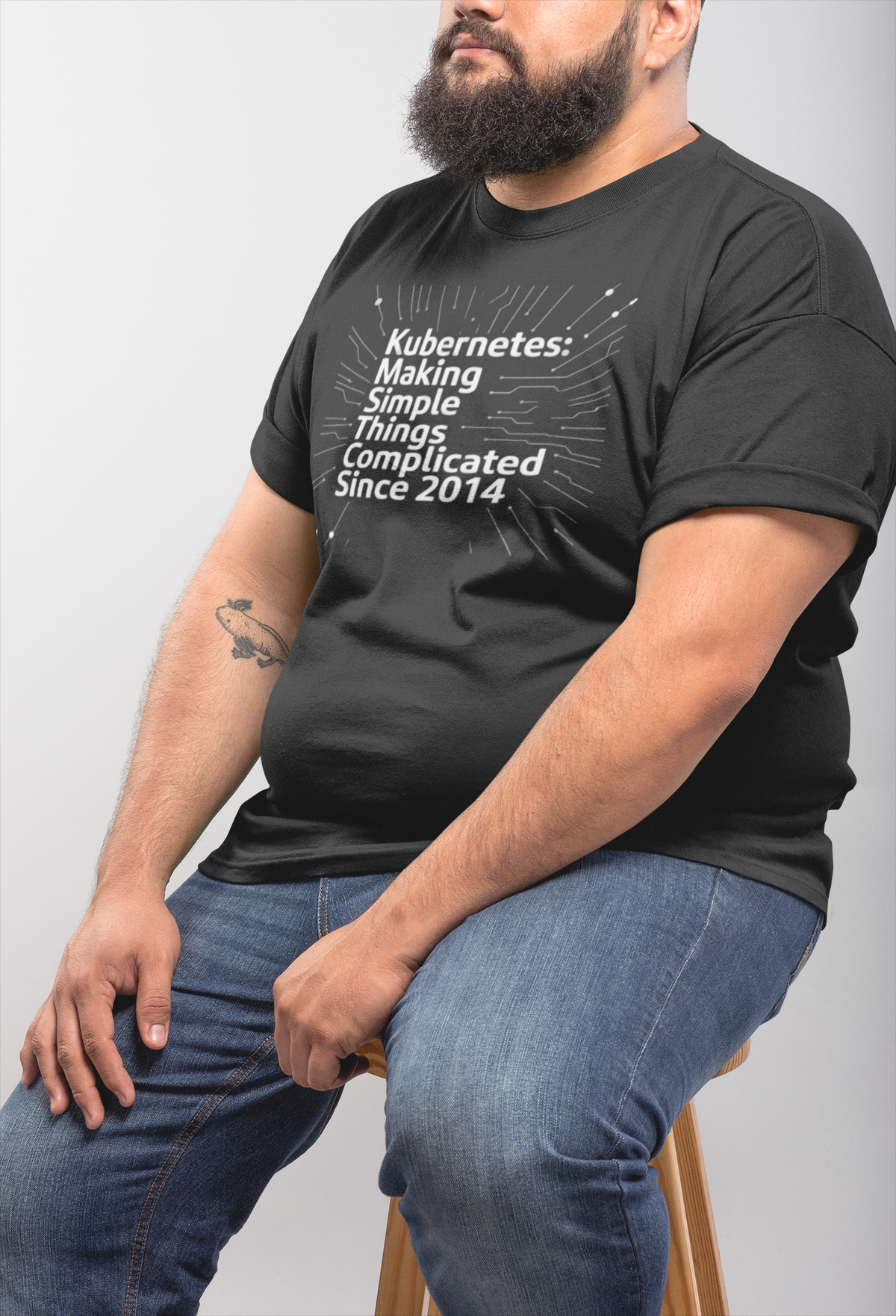 'Kubernetes: Making Simple Things Complicated Since 2014' t-shirt in charcoal, bearded man wearing it seated, DevOps gift