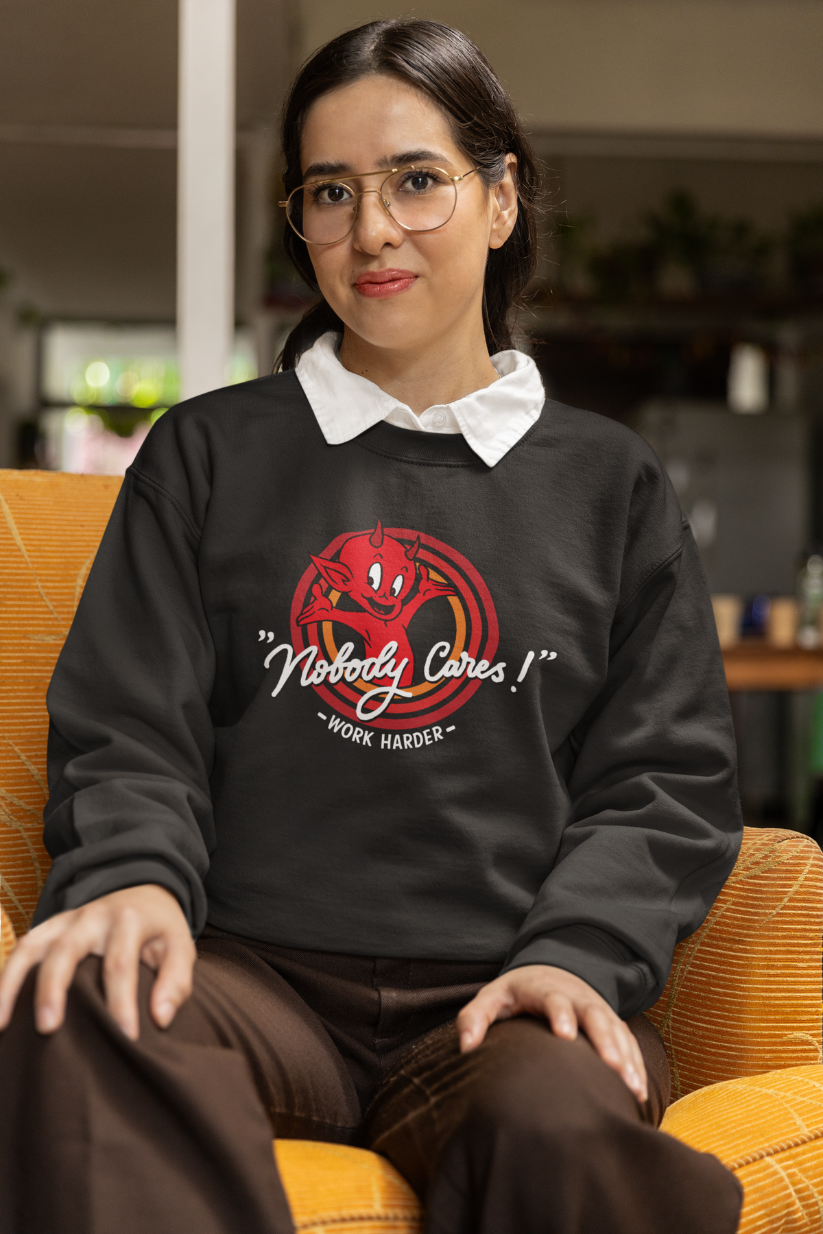 "Nobody Cares, Work Harder" devil graphic sweatshirt in black, woman wearing it seated in glasses, funny hustle gift