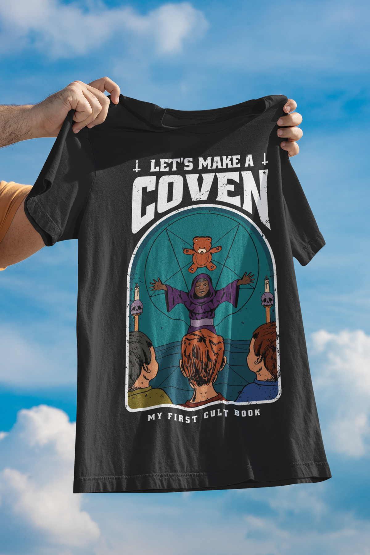'Let's Make A Coven' witch t-shirt in black, held up against a blue sky, spooky Halloween graphic