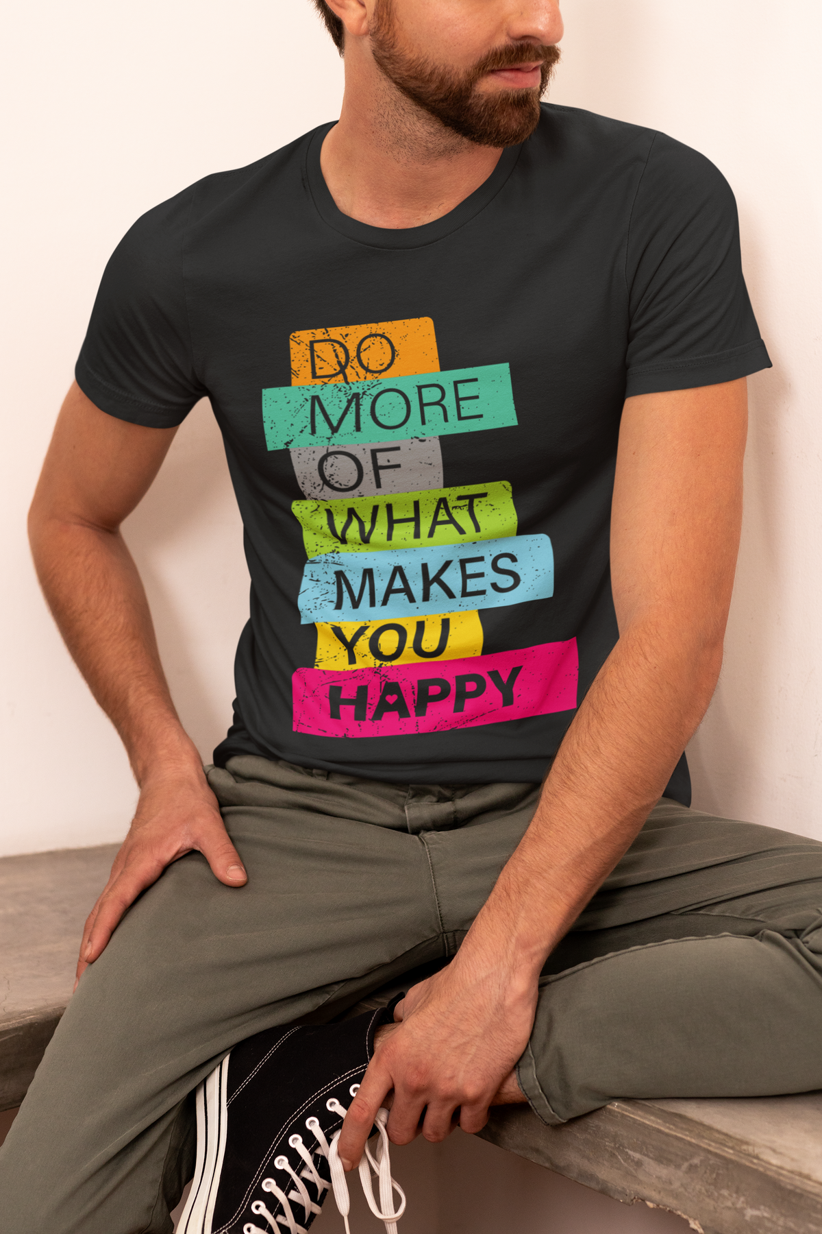 'Do More of What Makes You Happy' T-shirt in black, man sitting wearing it against a tan backdrop