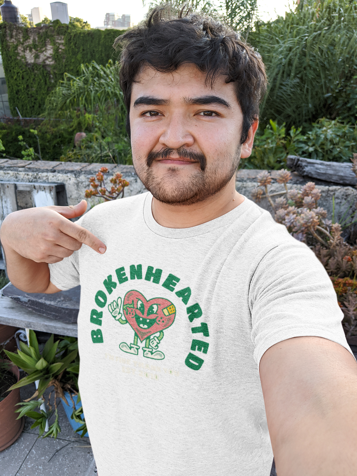 'Brokenhearted But Keeps On Rocking' t-shirt in white, man pointing at it outdoors, funny music gift