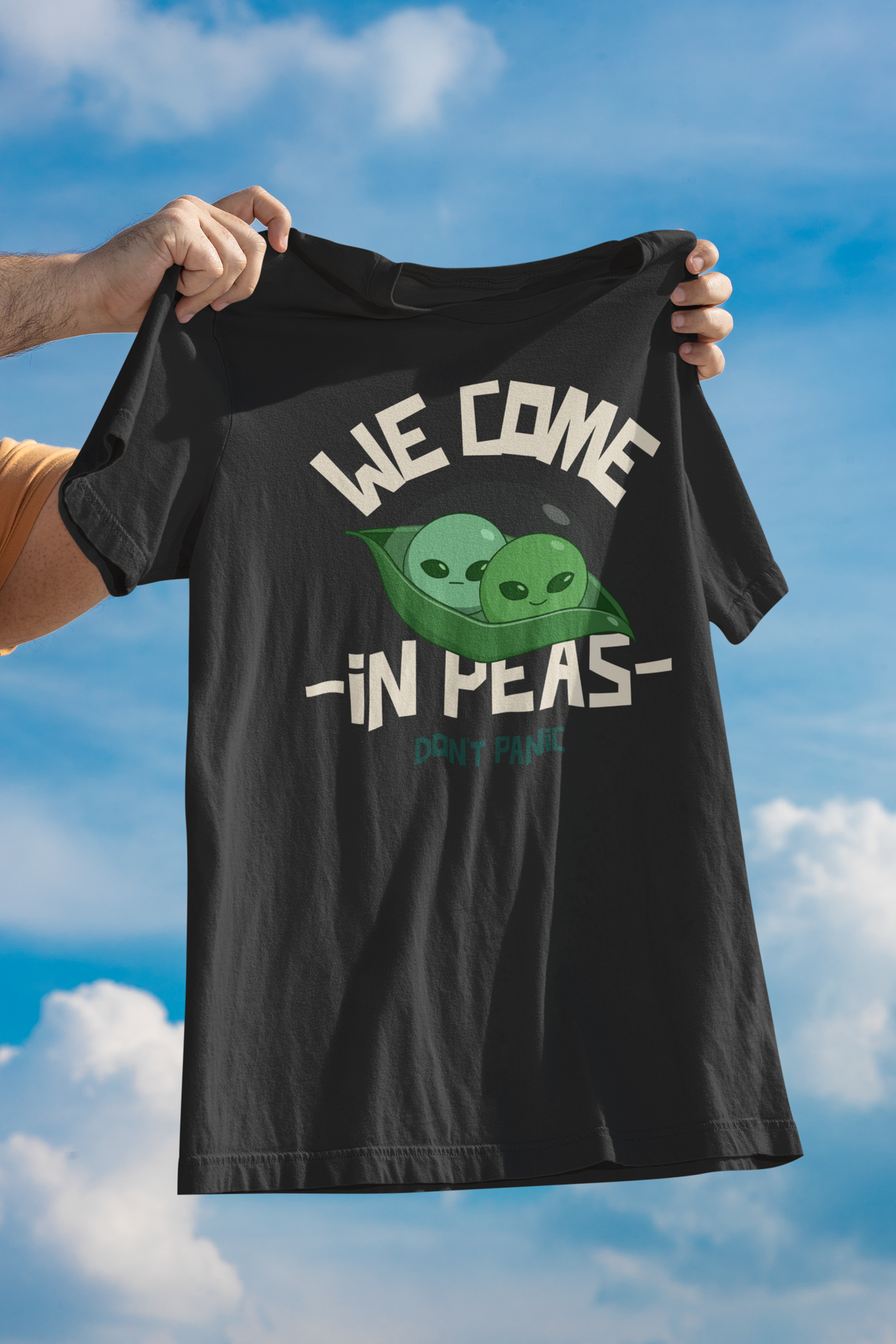 'We Come in Peas' t-shirt in black, person holding it up against a blue sky, funny alien pea pun tee gift