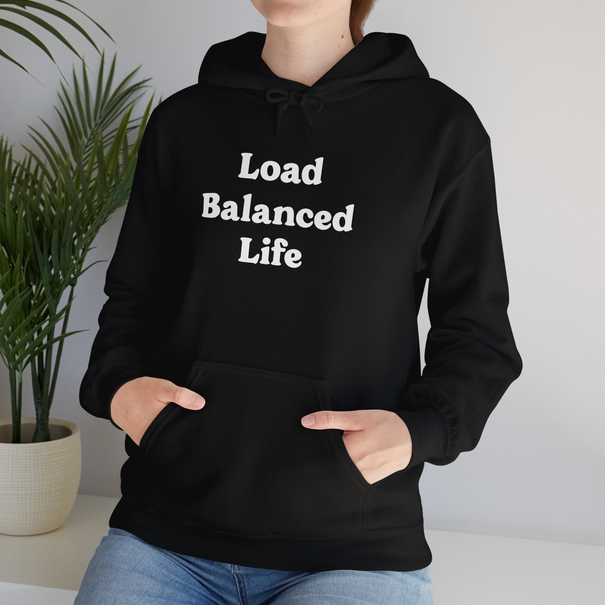 A person stands beside a potted plant, wearing the "Load Balanced Life" hooded sweatshirt. The seamless comfort of the design harmonizes beautifully with the serene atmosphere.