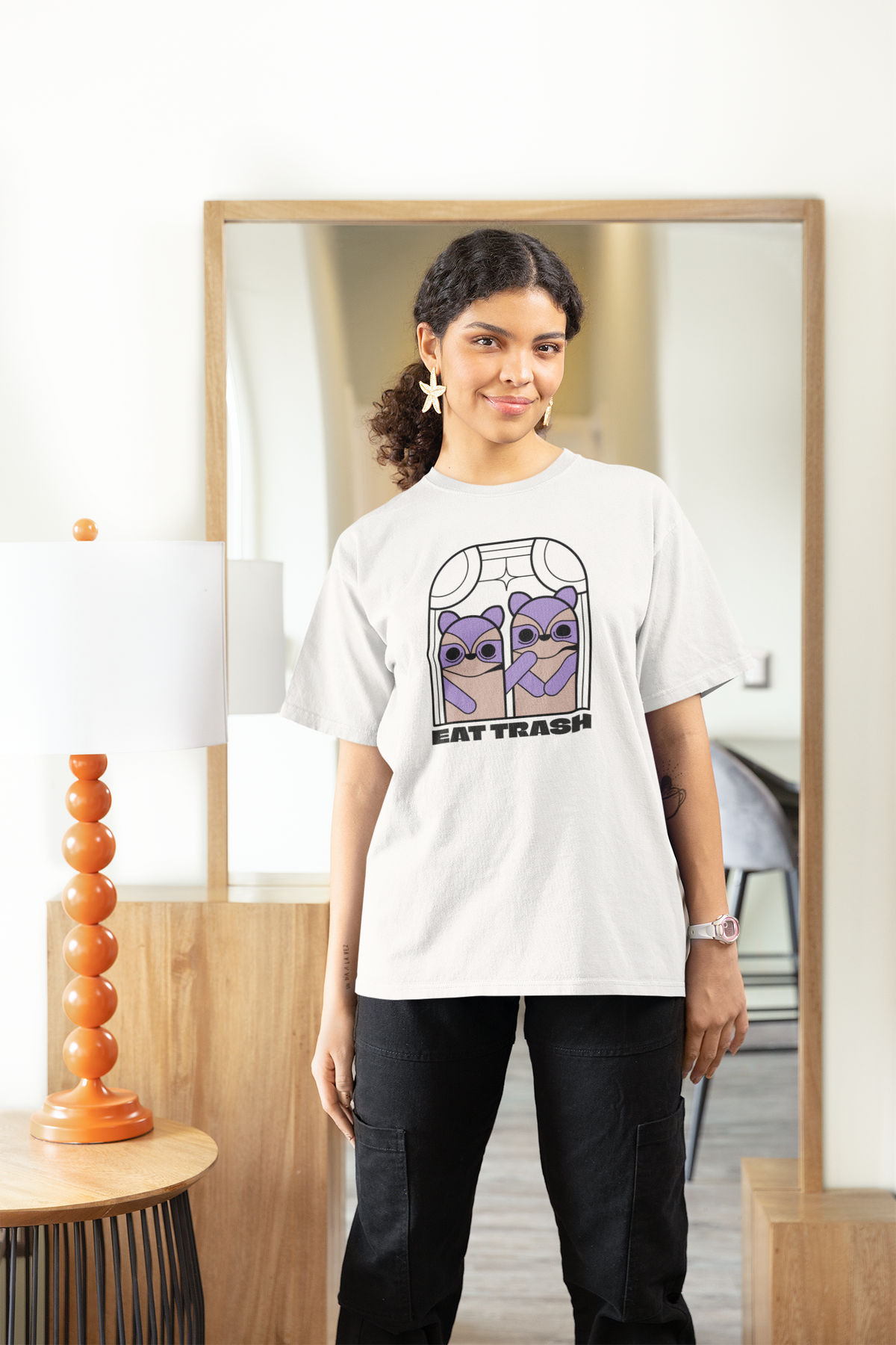 'Eat Trash' raccoon women's tee in white, woman wearing it in a stylish room, funny trash panda pun t-shirt