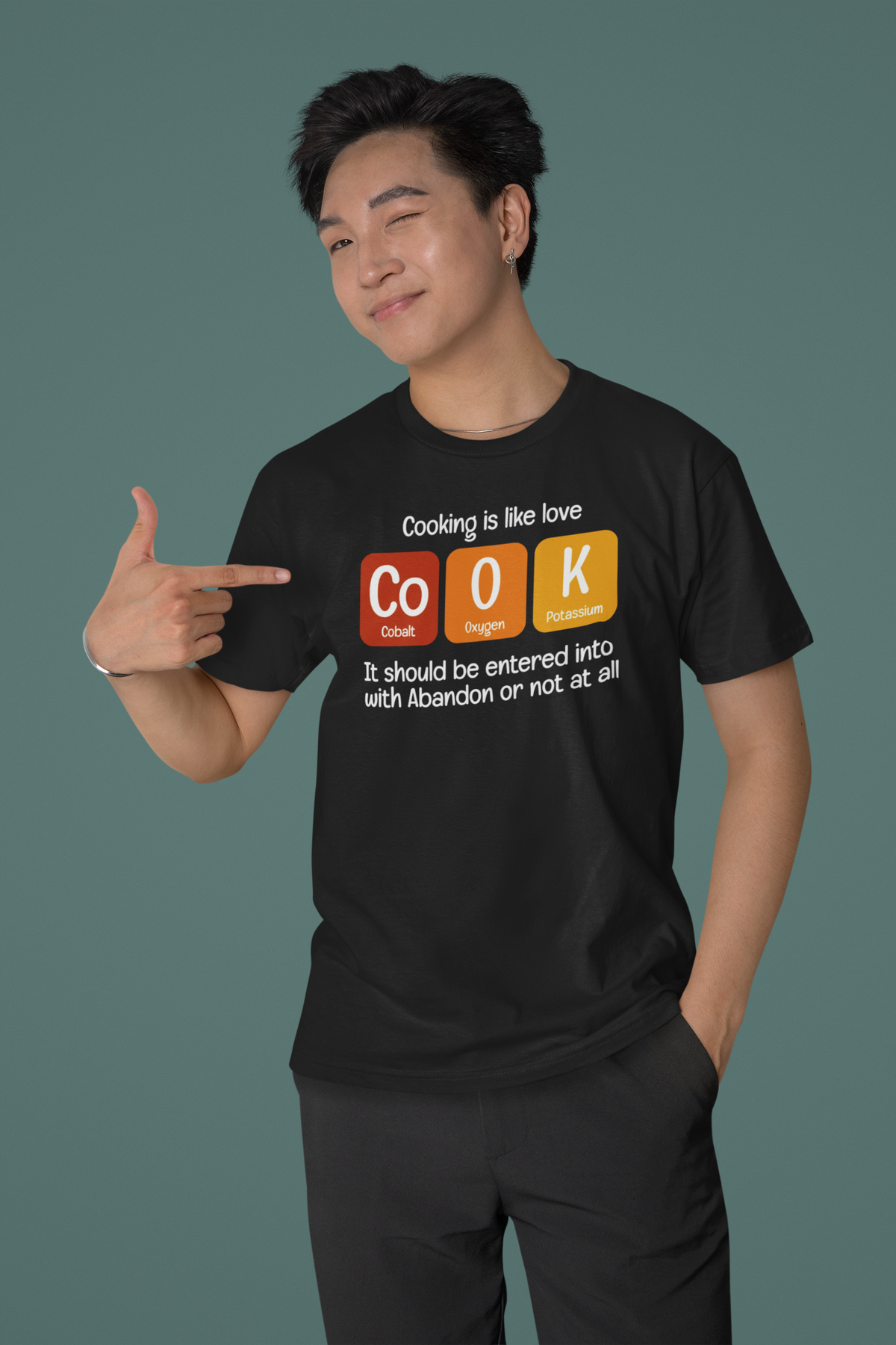 'Cook' periodic table t-shirt in black, man wearing it against green backdrop pointing at print, funny foodie gift