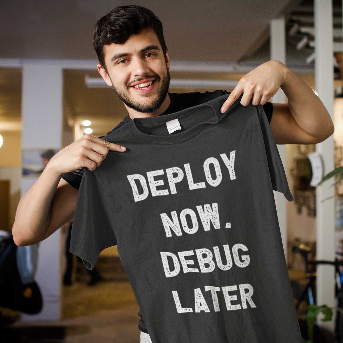 'Deploy Now, Debug Later' t-shirt in heather charcoal, man holding it up smiling, funny gift for coders