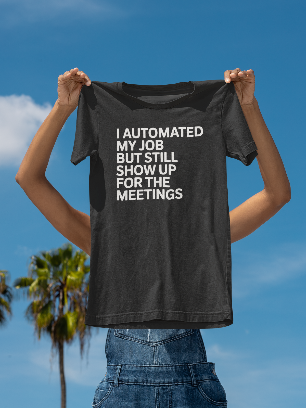 'I Automated My Job But Still Show Up for the Meetings' t-shirt in black, woman holding it up outdoors, funny developer gift