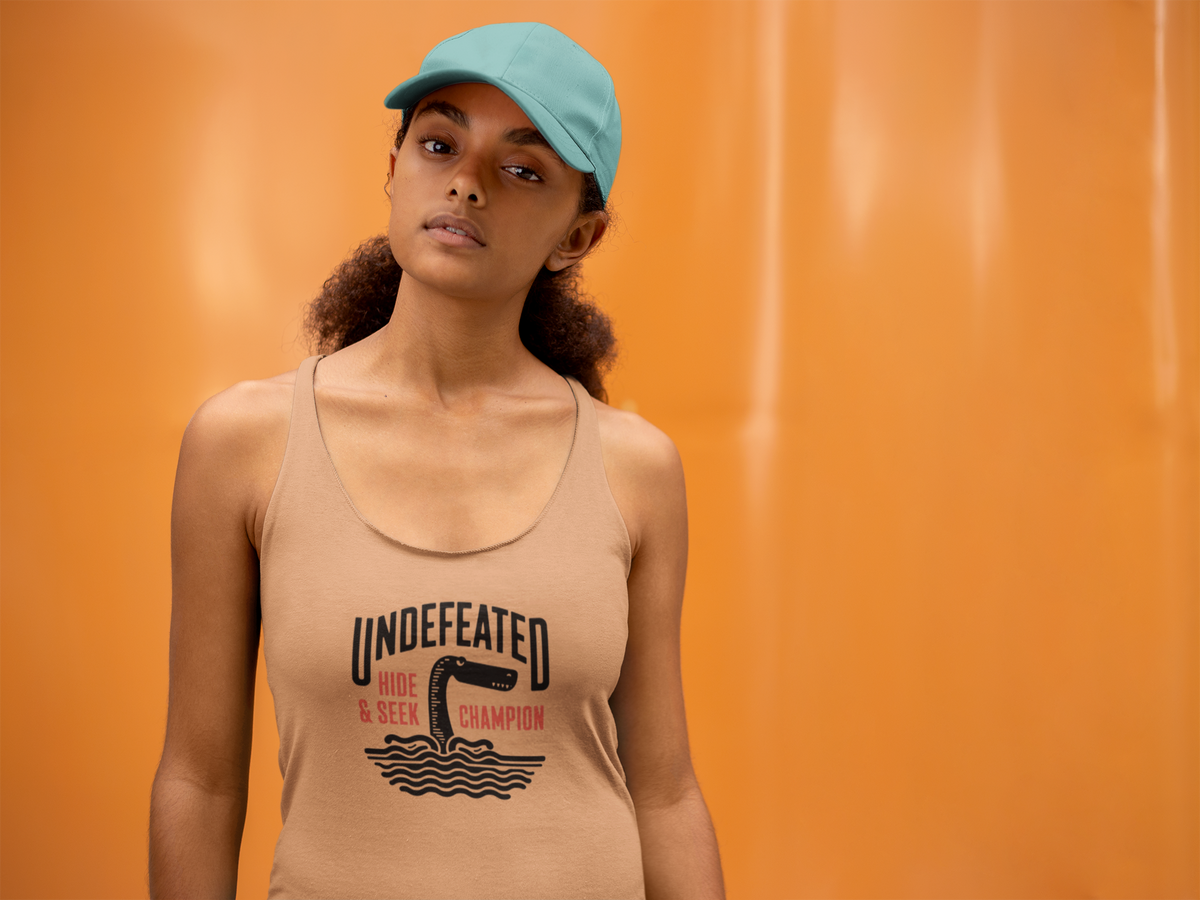 'Undefeated Hide and Seek Women's Tank' in light orange, woman by an orange wall, Tank Top mockup, Geekpsters