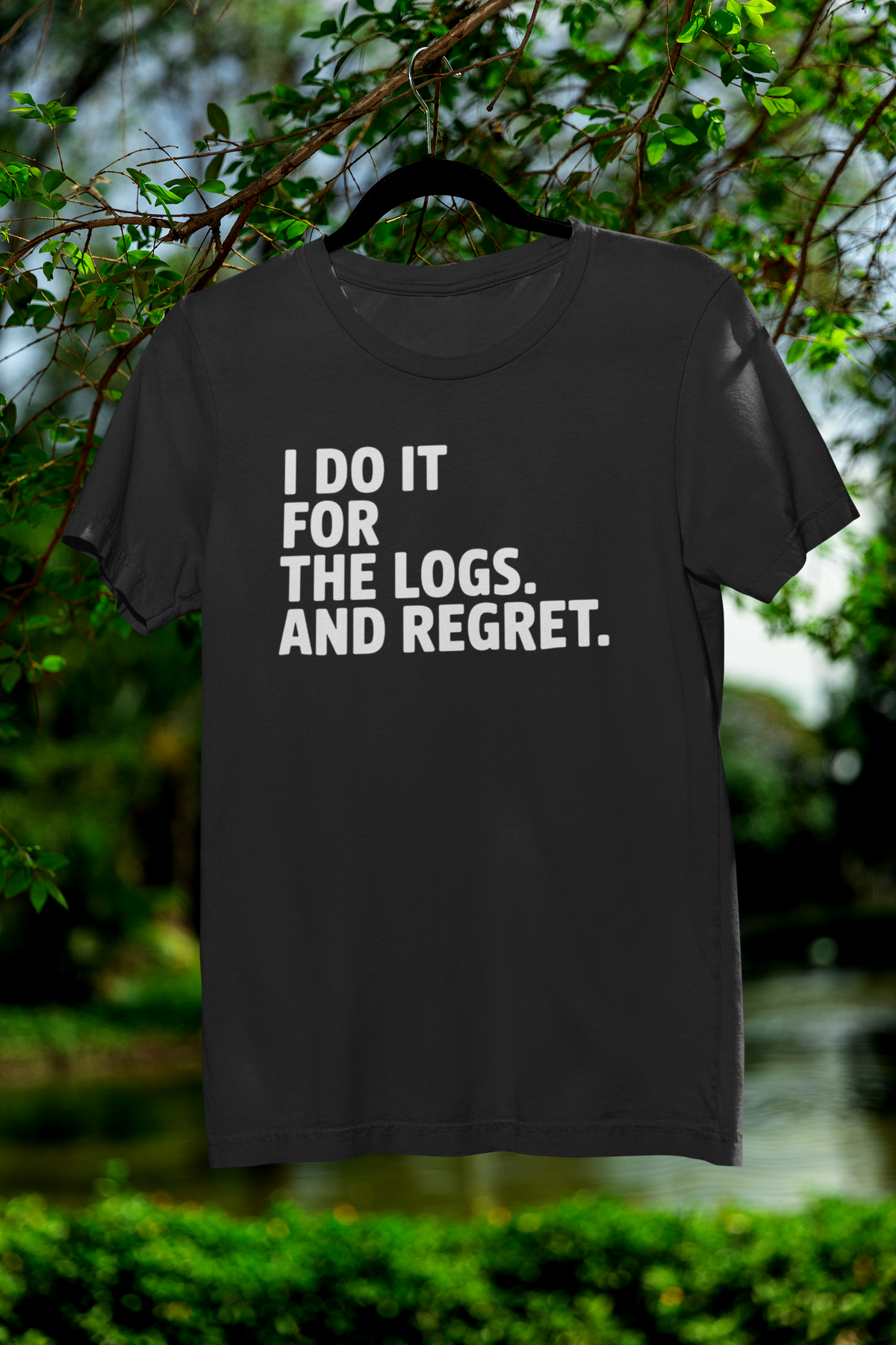 'I Do It For The Logs. And Regret.' t-shirt in black, hanging outdoors among green leaves, funny programmer shirt