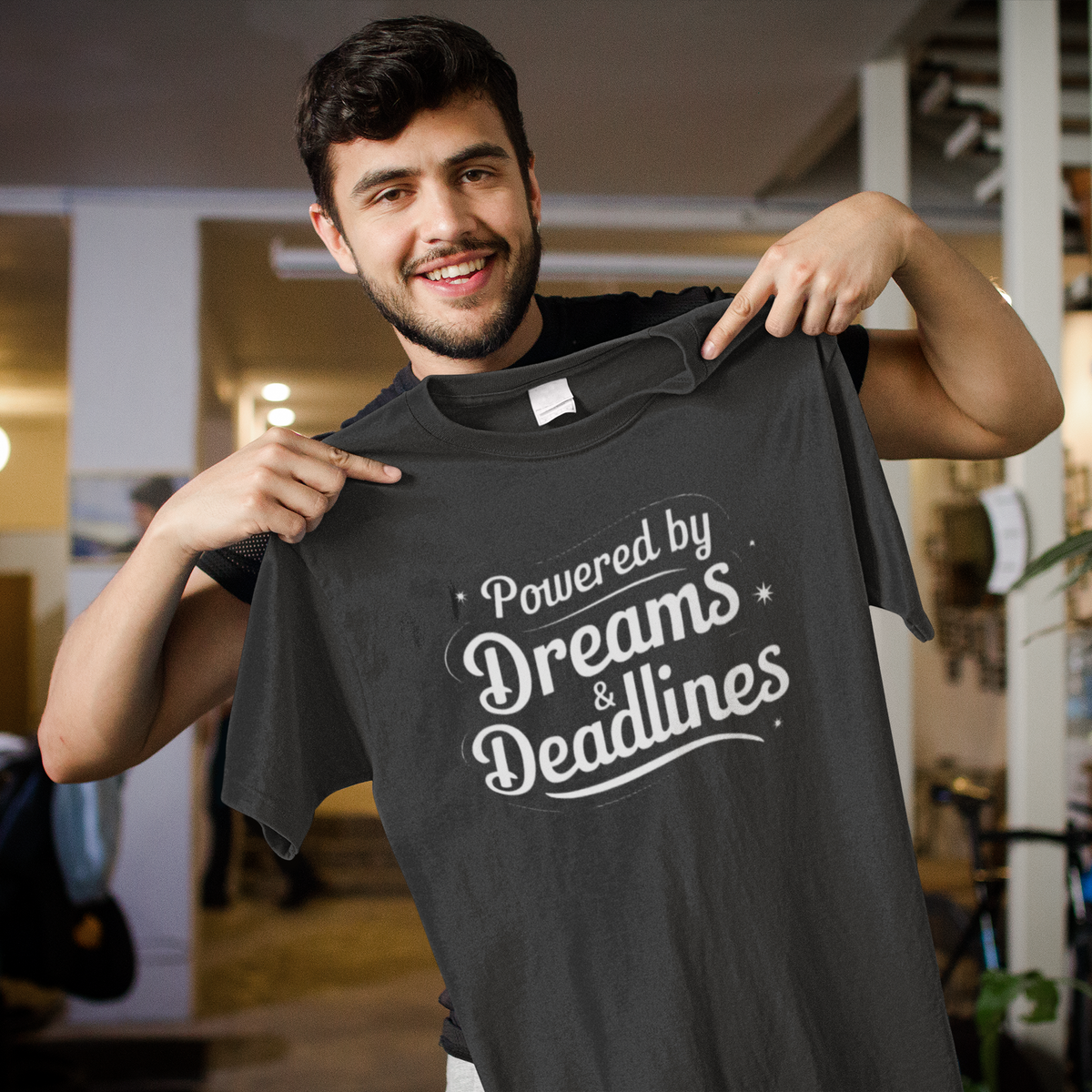 'Powered by Dreams & Deadlines' organic t-shirt in black, man holding it up indoors, funny developer gift