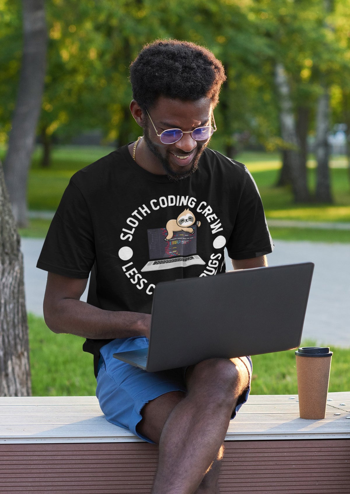 Sloth Coding Crew tee in black, man coding on a laptop on a park bench, funny developer gift