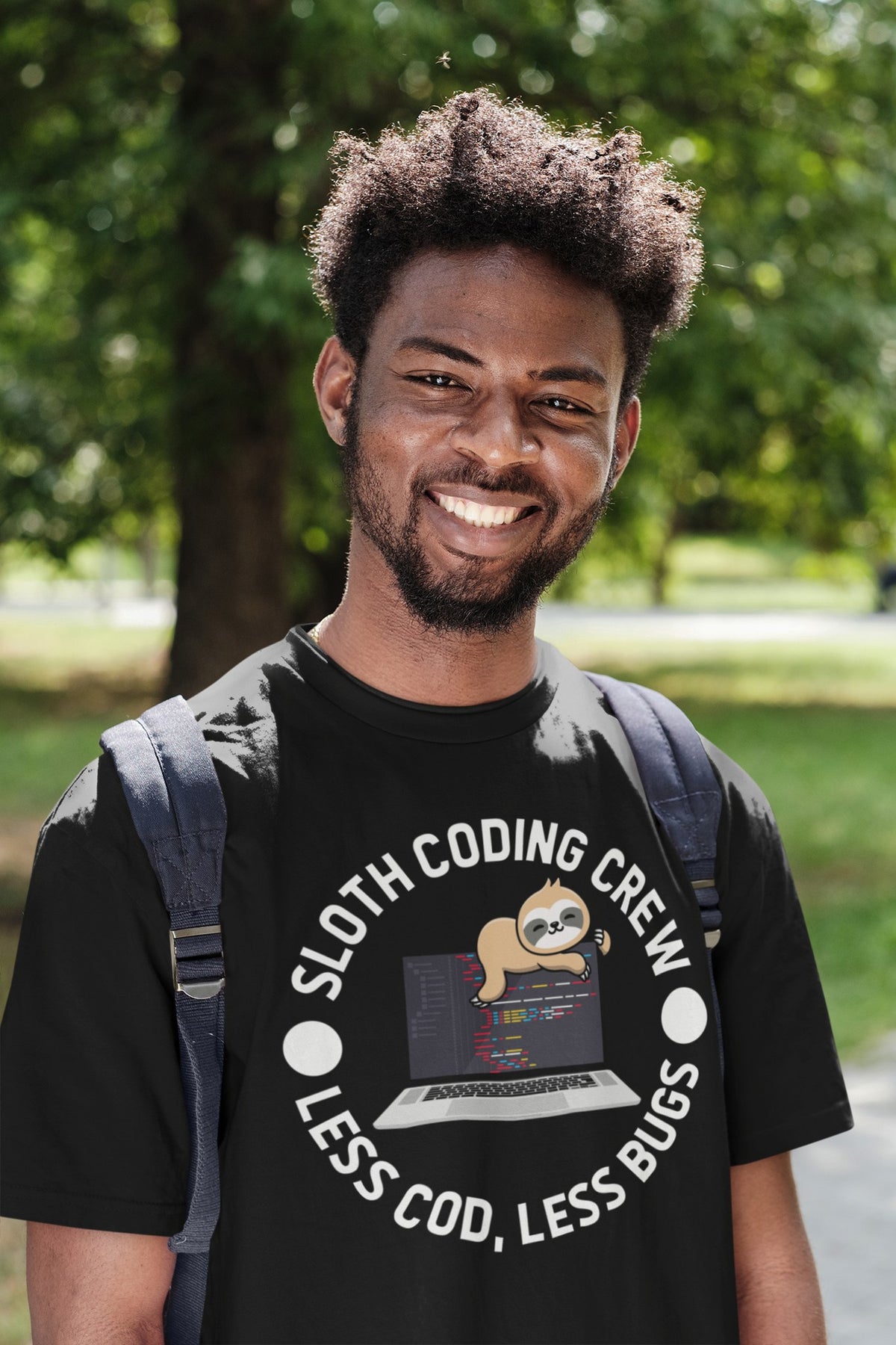 Sloth Coding Crew tee in black, smiling man with a backpack in a sunny park, programmer gift