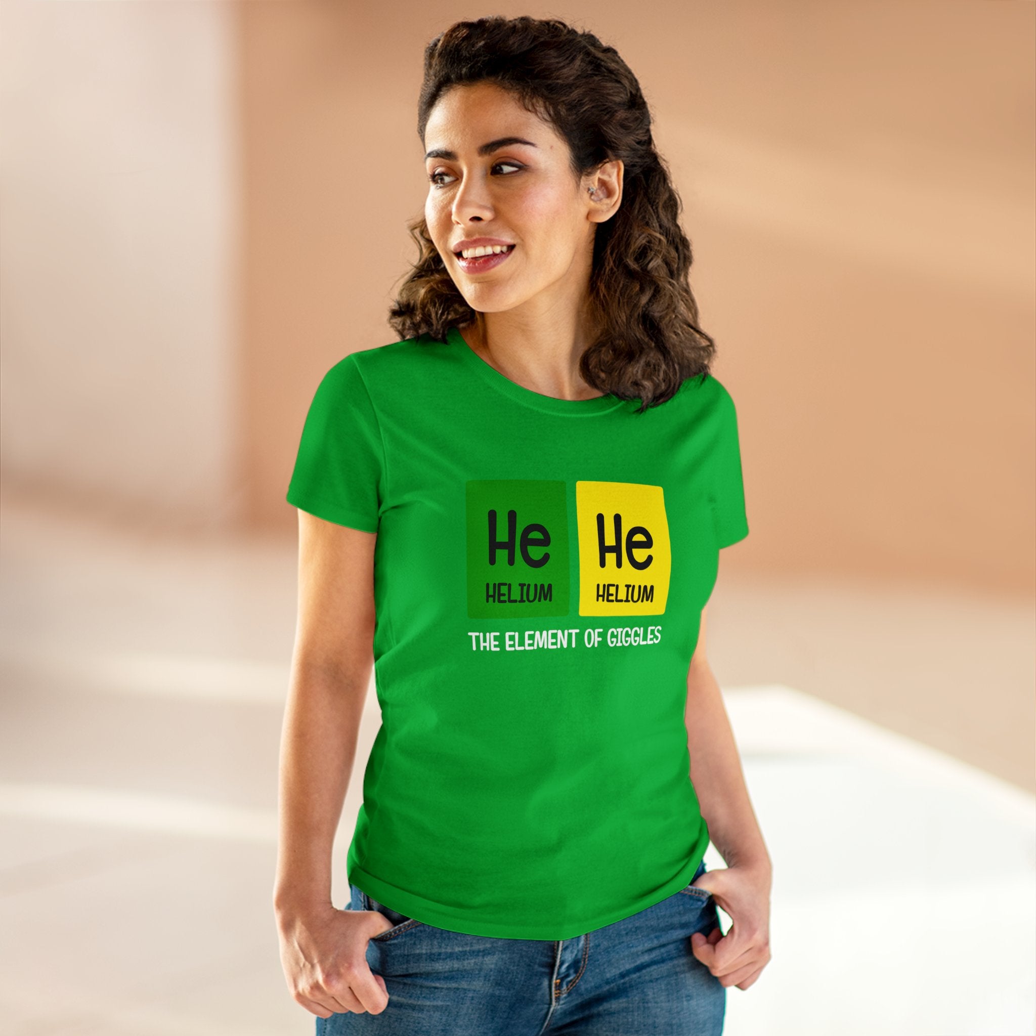 A person wearing an ultra-cozy He-He - Women'sTee made from ethically grown US cotton, adorned with helium elements and the text "The element of giggles," stands indoors smiling.