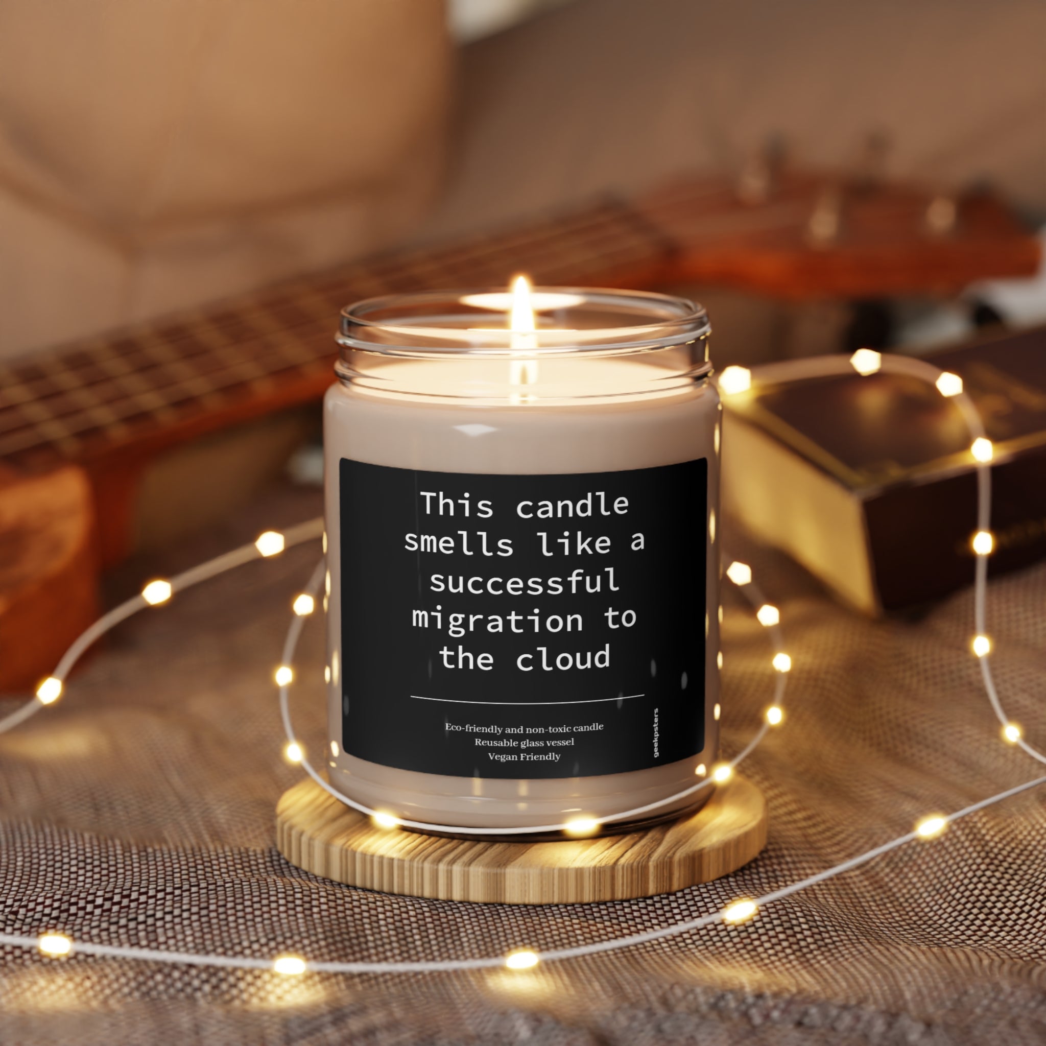 A lit "This candle smells like a successful migration to the cloud" scented soy wax candle with a humorous label related to cloud technology on a wooden surface surrounded by fairy lights.