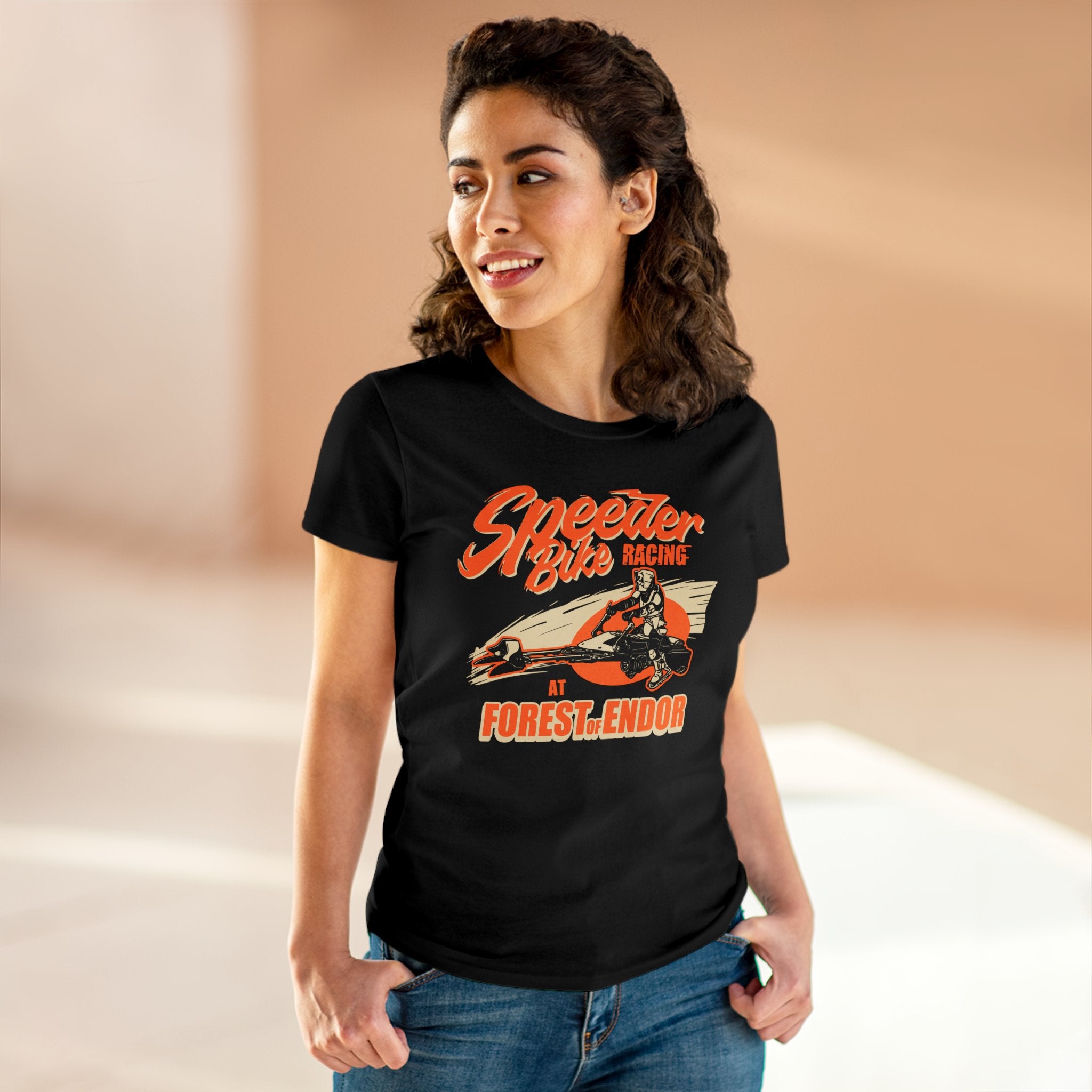 A person stands indoors, smiling and wearing a pre-shrunk cotton black t-shirt that reads "Speeder Bike Racing - Women's Tee" with an image of a person riding a speeder bike.