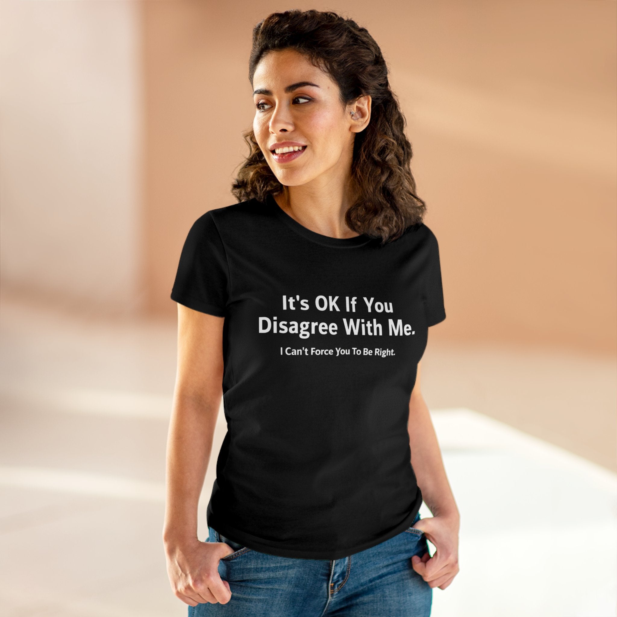 A woman wearing a black t-shirt with the empowering print "It's OK If You Disagree With Me. I Can't Force You To Be Right." is standing and smiling. This pre-shrunk "It's Ok If You Disagree With Me - Women's Tee" epitomizes confidence and wit, perfect for making a bold statement.