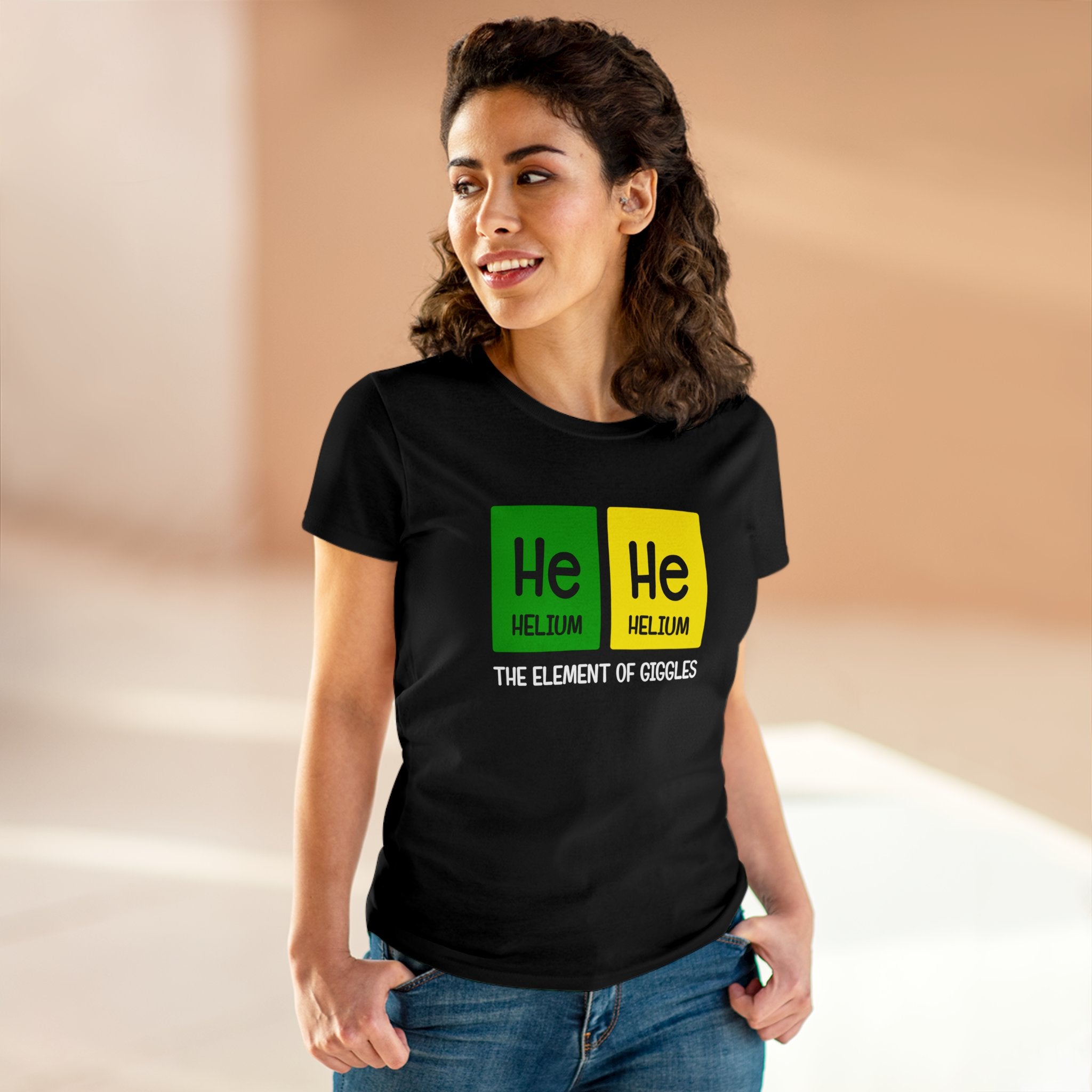 A woman wearing a black T-shirt with a periodic table joke that reads "He-He - Women'sTee" stands indoors with hands in her ethically grown cotton jeans pockets, looking to her right and smiling.