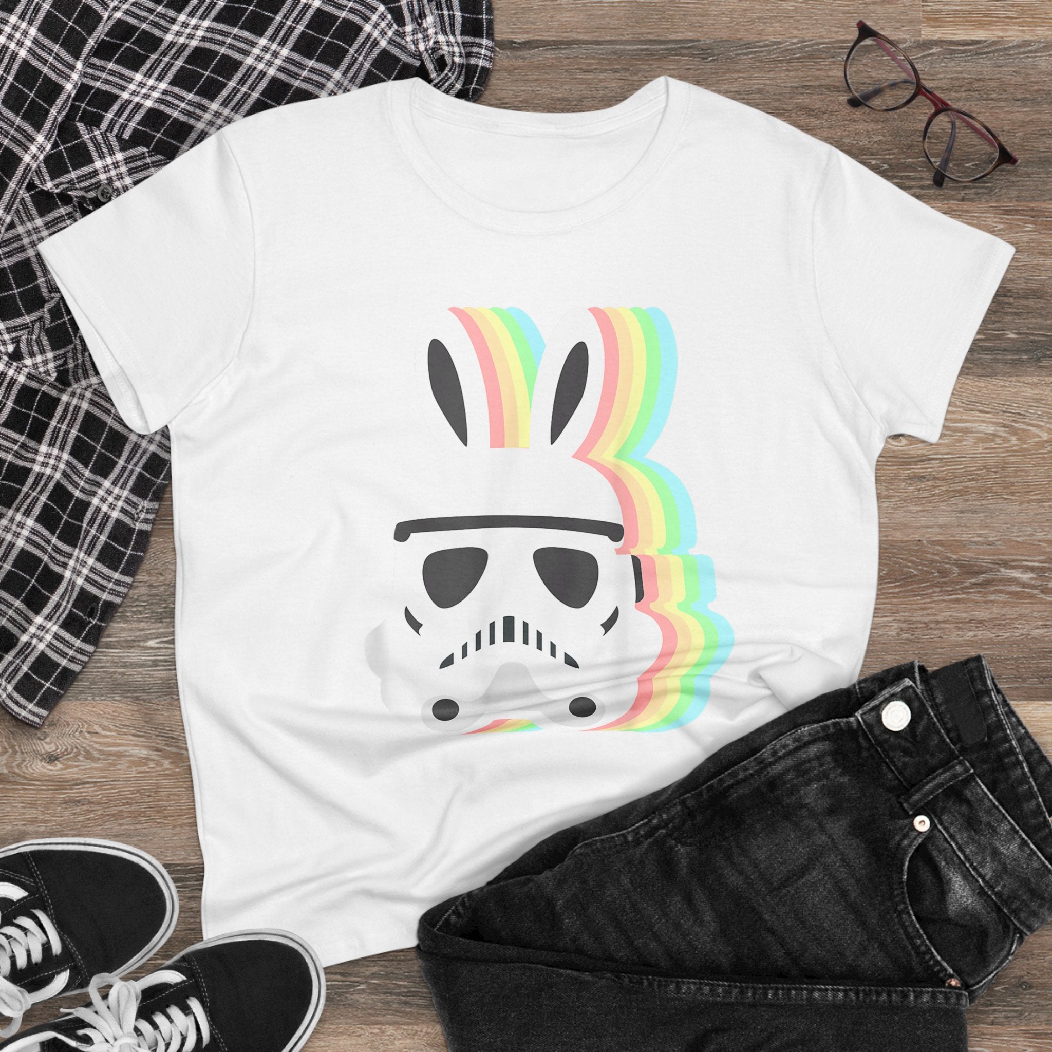 A Star Wars Easter Stormtrooper - Women's Tee featuring an Easter Stormtrooper helmet with bunny ears and rainbow stripes. The white t-shirt is laid on a wooden surface next to black jeans, a plaid shirt, and a pair of glasses. Perfect for Star Wars fans with a playful twist!