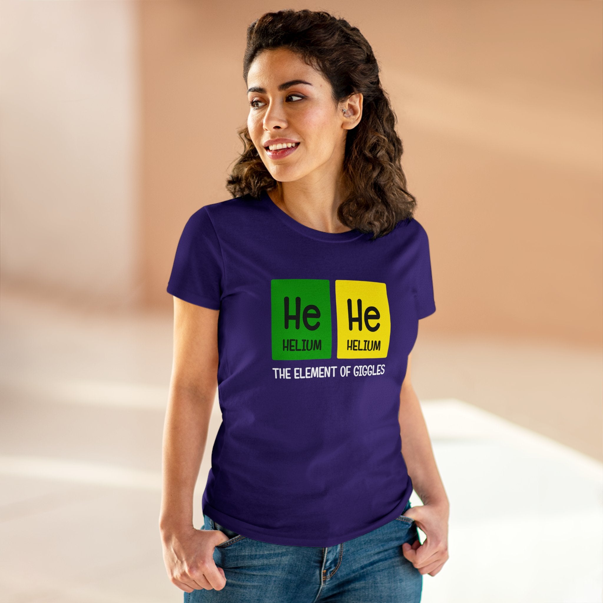 A person wearing a chic and comfy purple He-He - Women'sTee with the periodic table symbols for Helium (He) and the text "He He" and "The element of giggles," standing indoors with hands in pockets. The shirt is crafted from ethically grown US cotton.