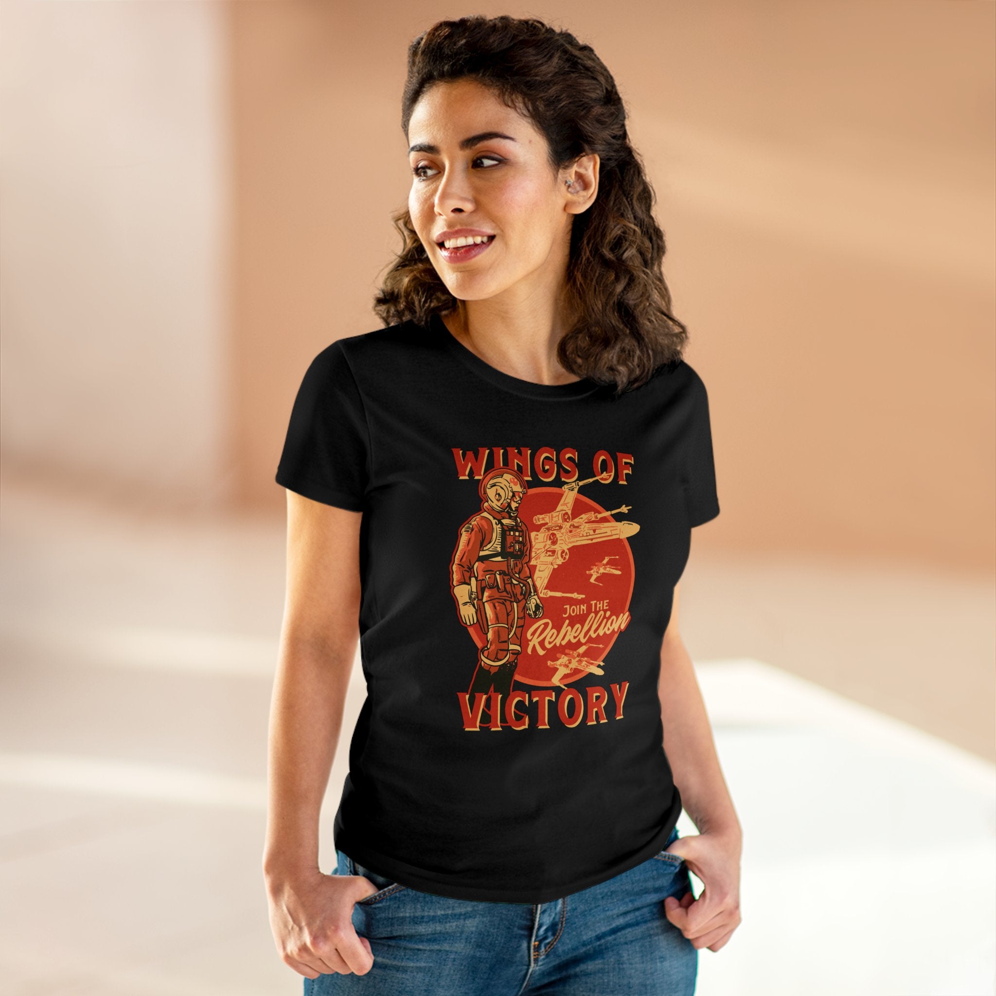 A woman wearing a black, pre-shrunk Wings of Victory - Women's Tee with a graphic design featuring a character, text that reads "Wings of Victory" and "Join the Rebellion," and jeans, smiles while standing indoors.