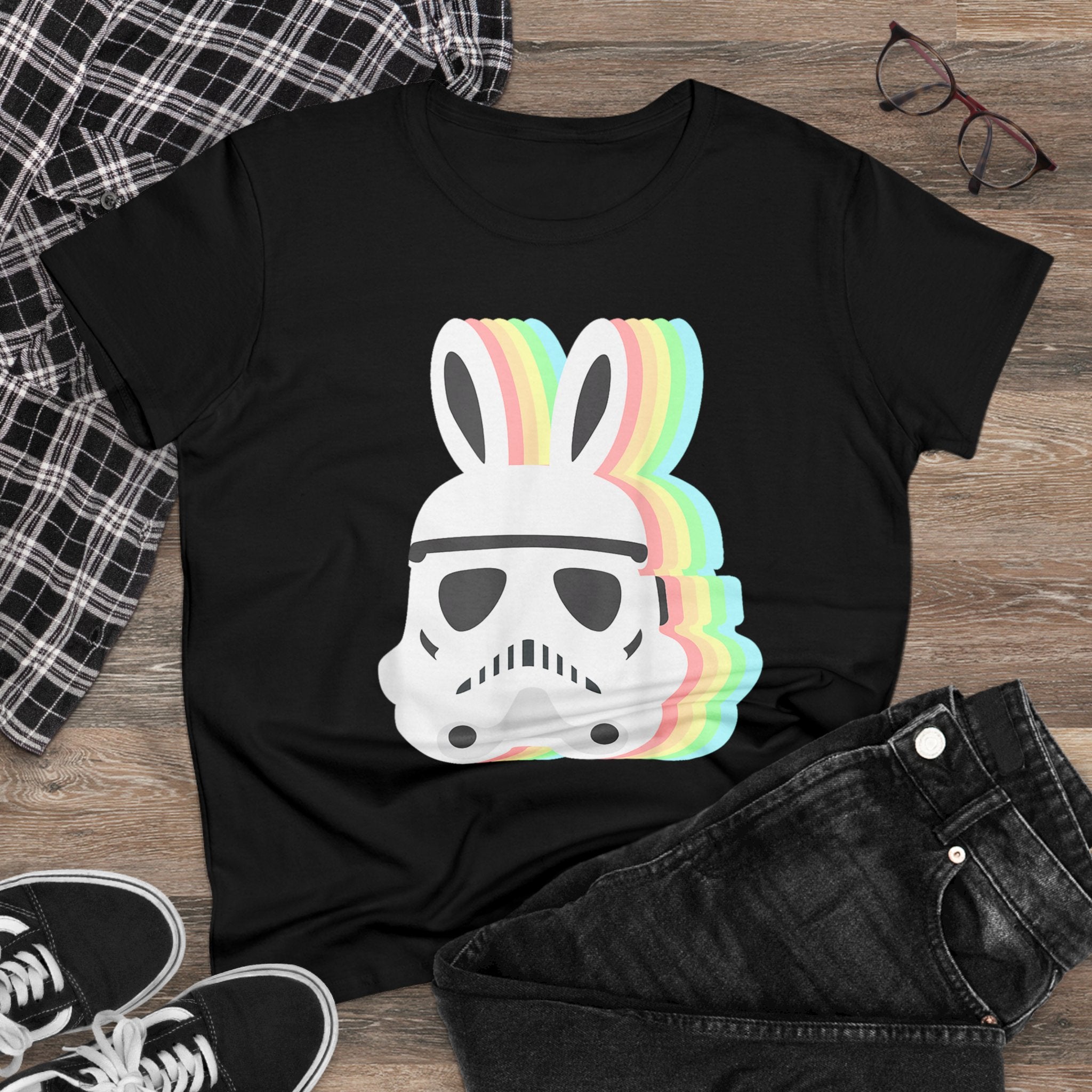 Black Star Wars Easter Stormtrooper - Women's Tee featuring a graphic of an Easter Stormtrooper helmet with bunny ears and rainbow layers. Shirt lies on a wooden floor, accompanied by plaid shirt, black jeans, sneakers, and eyeglasses.