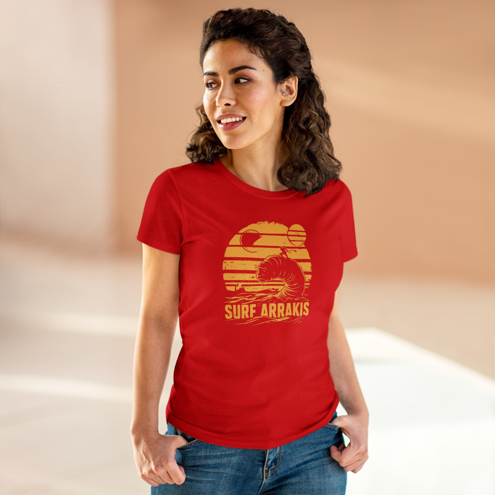 A woman with dark hair stands indoors wearing a red "Surf Arrakis - Women's Tee" made from soft light cotton and blue jeans against a blurred background.