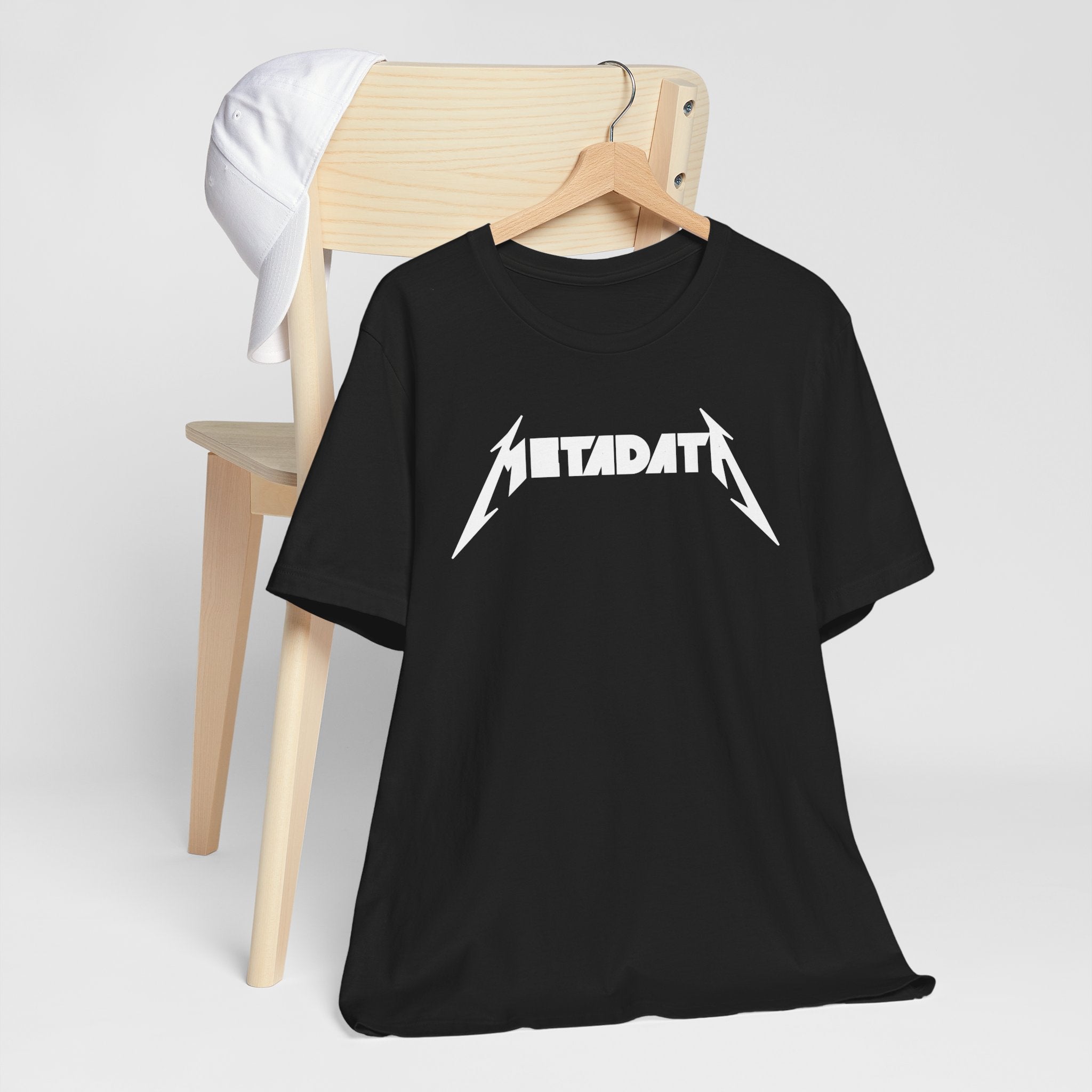 The "Metadata" T-shirt, crafted from Airlume combed cotton and showcasing a Metallica-style font design, artfully drapes over a wooden chair. The display is enhanced by a white cap hanging on the chair's backrest, offering supreme comfort.