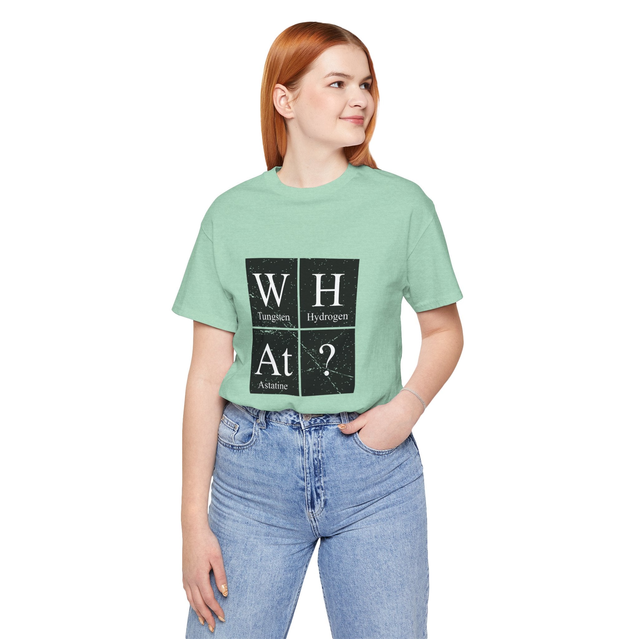 Woman in a green unisex jersey tee with W-H-At-? design, standing with hands in jeans pockets.