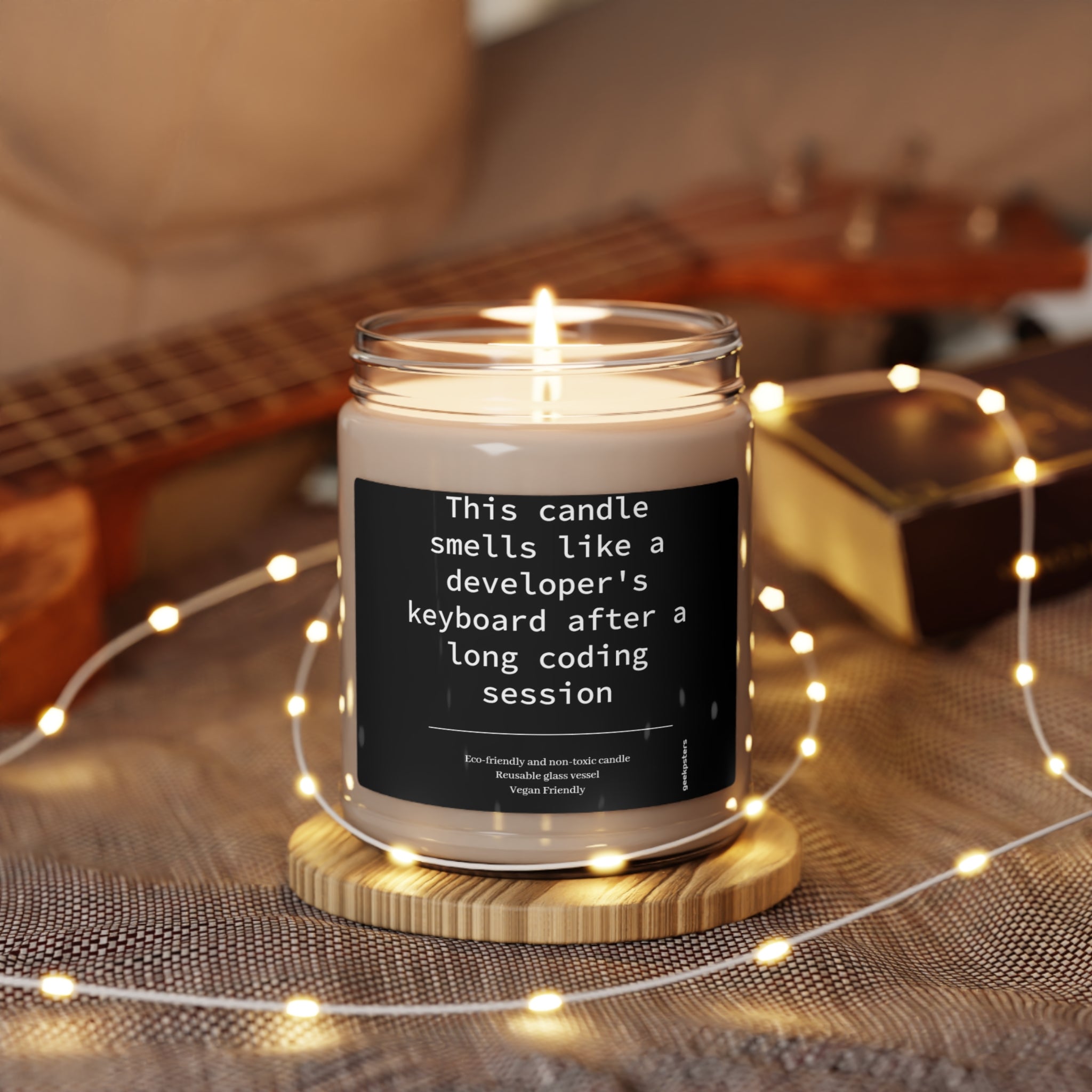 A lit scented candle with a natural soy wax blend and a humorous label suggesting it smells like the scented soy candle, 9oz, placed among fairy lights and gift boxes.